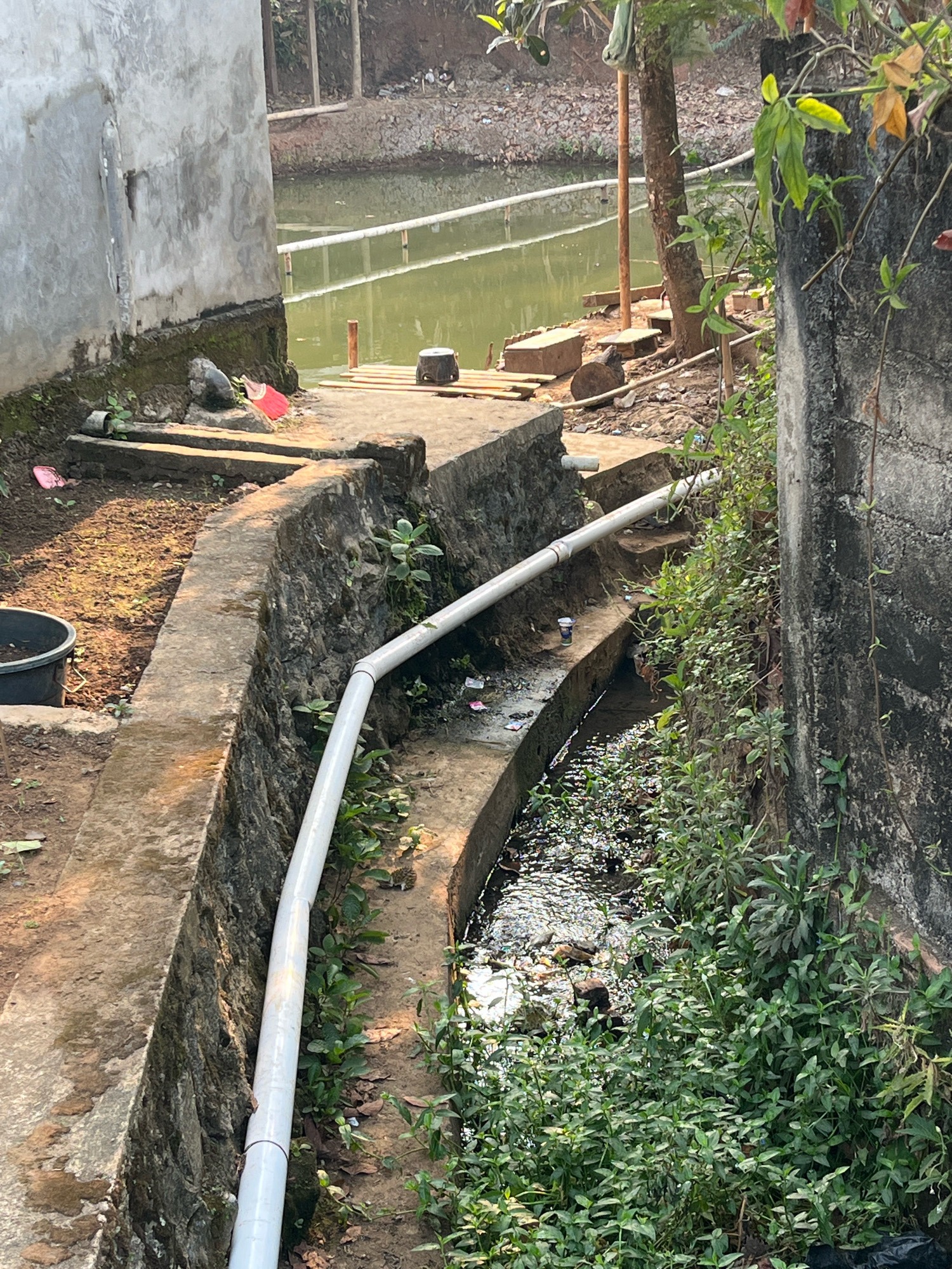In Margodadi, Indonesia, a concrete drainage channel runs alongside a murky, green body of water. A white PVC pipe extends diagonally across the scene, supported by the channel's concrete walls. Vegetation grows thickly along the sides of the channel and on the surrounding earth. A concrete building is visible in the background. The area appears to be an outdoor, undeveloped space, possibly on the outskirts of a residential or industrial zone.