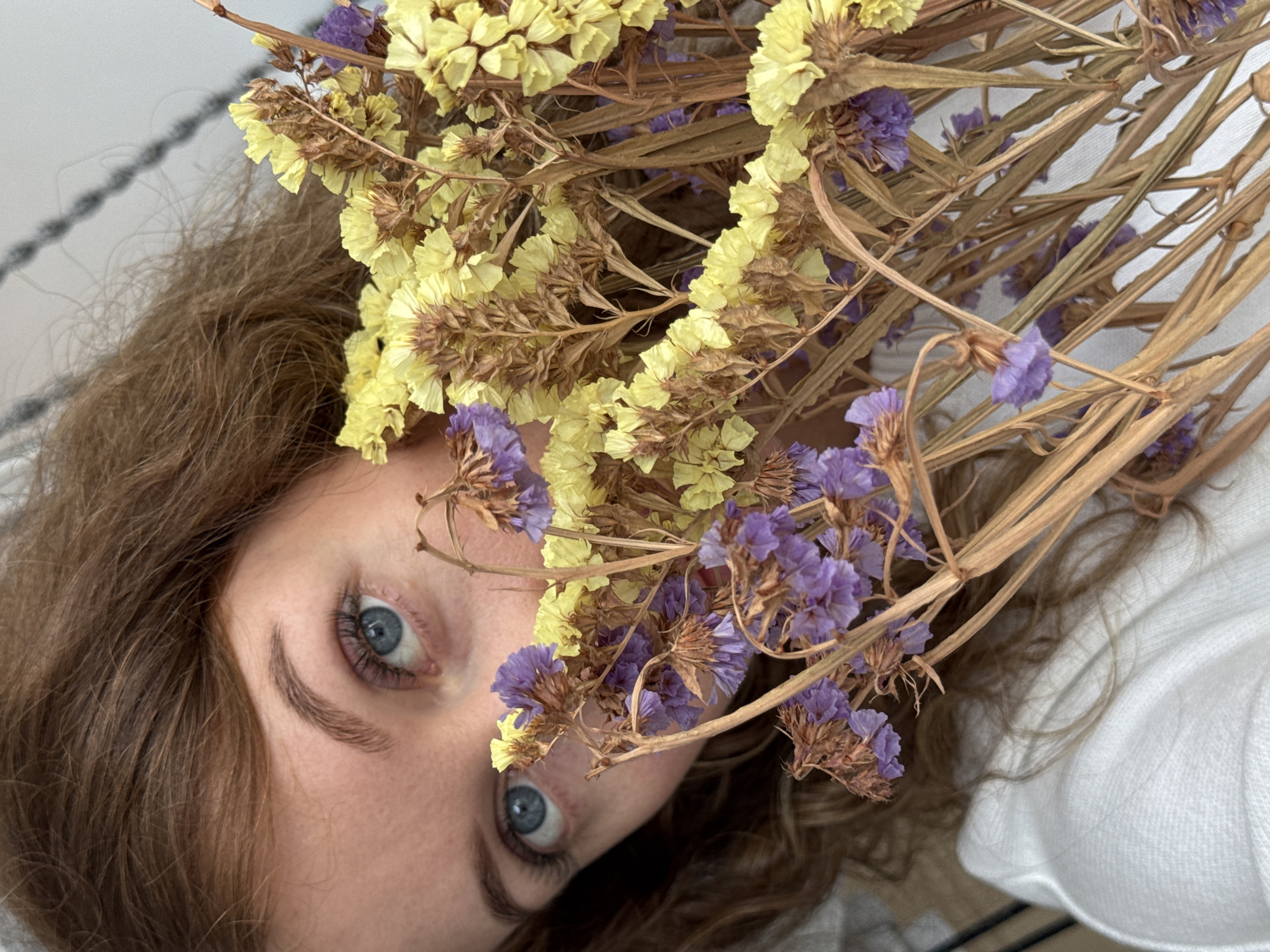A woman is pictured from an angled close-up perspective, with the image rotated. She has light skin, brown hair, and blue eyes. A bouquet of dried flowers, featuring yellow and purple blooms, is arranged in front of her face. The woman is wearing a white garment. The location is not identifiable as Chaikovsky, Russia.
