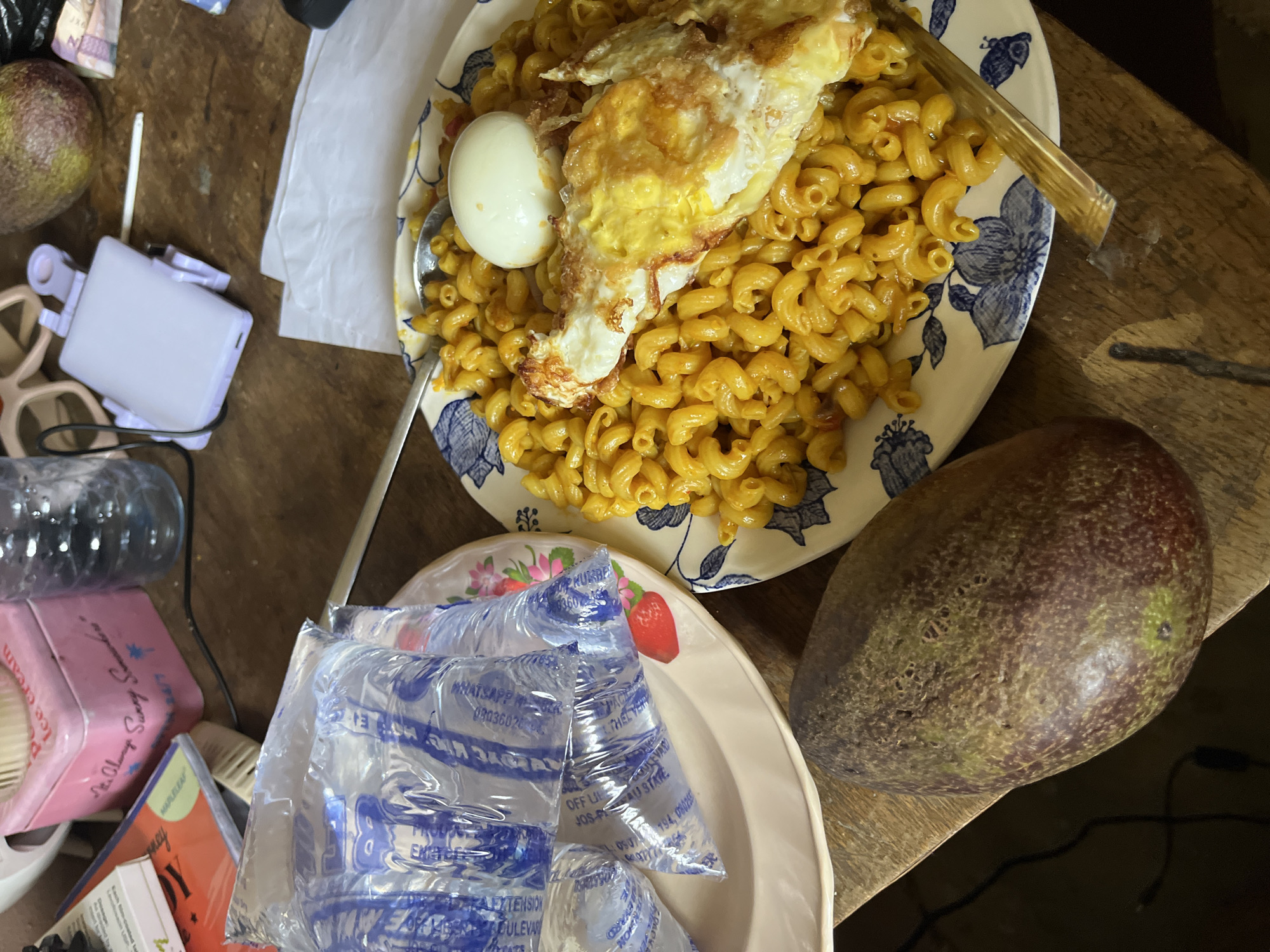 The image shows a wooden table with a plate of macaroni and cheese, a fried egg, and a hard-boiled egg. There are also several packets of water, an avocado, and a pink tin with the words "It's Always Sunny Somewhere" written on it. A glass bottle and some books are also visible on the table. The scene appears to be an outdoor setting, possibly a market or roadside stall, given the presence of the water packets and the rustic wooden table. The avocado and the style of the food suggest a West African context, possibly Nigeria, given the prompt's reference to Rafin Par. However, without more specific environmental cues, it is difficult to pinpoint the exact location within Rafin Par.