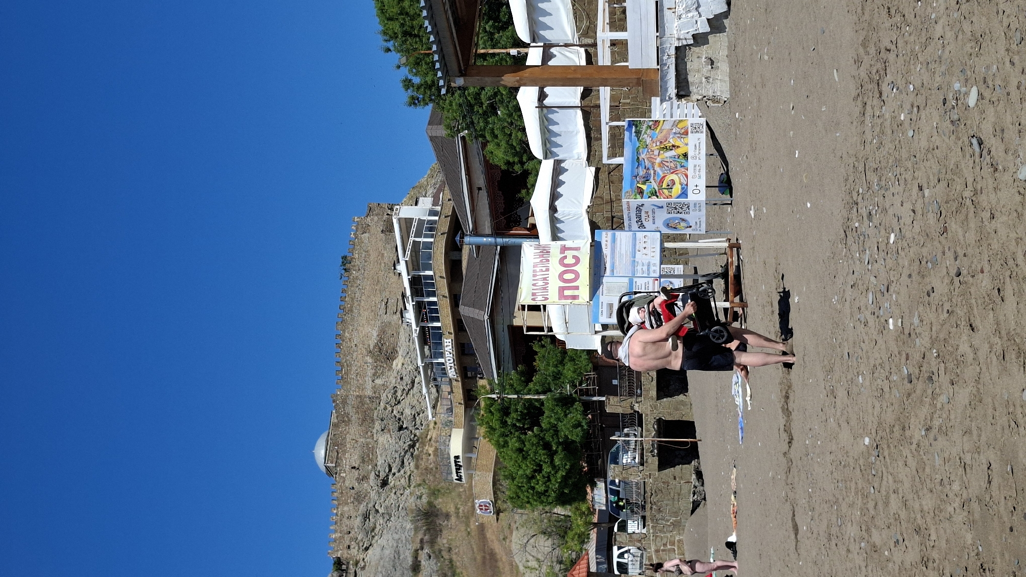 This image depicts a sunny beach scene with a man, buildings, and a fortified hill under a clear blue sky. The photo is rotated 90 degrees clockwise, showing the sky on the left and the beach on the right if viewed incorrectly. When correctly oriented, the horizon is level.
In the foreground, a shirtless man wearing dark shorts sits on a small black and red recreational vehicle, possibly a quad bike or a utility cart, facing slightly away from the camera. The ground beneath him is a sandy and pebbly beach.
Behind the man, several informational signs are visible. A prominent yellow sign reads "СПАСАТЕЛЬНЫЙ ПОСТ" (Lifeguard Post) in red. Other signs advertise "Аквапарк СУДАК" (Aquapark Sudak) with an address "ул. Паприка, 77" and a phone number "567-94-77", along with a "0+" age rating and QR codes. White beach canopies or awnings are arranged further back, likely covering seating areas.
Behind these structures, several multi-story buildings with balconies and large windows line the beachfront. One building clearly displays "РЕСТОРАН" (Restaurant) and "АСТАРТА" (Astarta) on its facade. Green trees are interspersed among the buildings. To the left, a large, rocky hill or cliff rises, topped by an ancient-looking fortified wall with battlements, suggesting a historical castle or fortress overlooking the area. The sky above is a brilliant, cloudless blue. The scene captures a vibrant summer day at a coastal resort, likely in Sudak, Crimea, as indicated by the aquapark advertisement.
