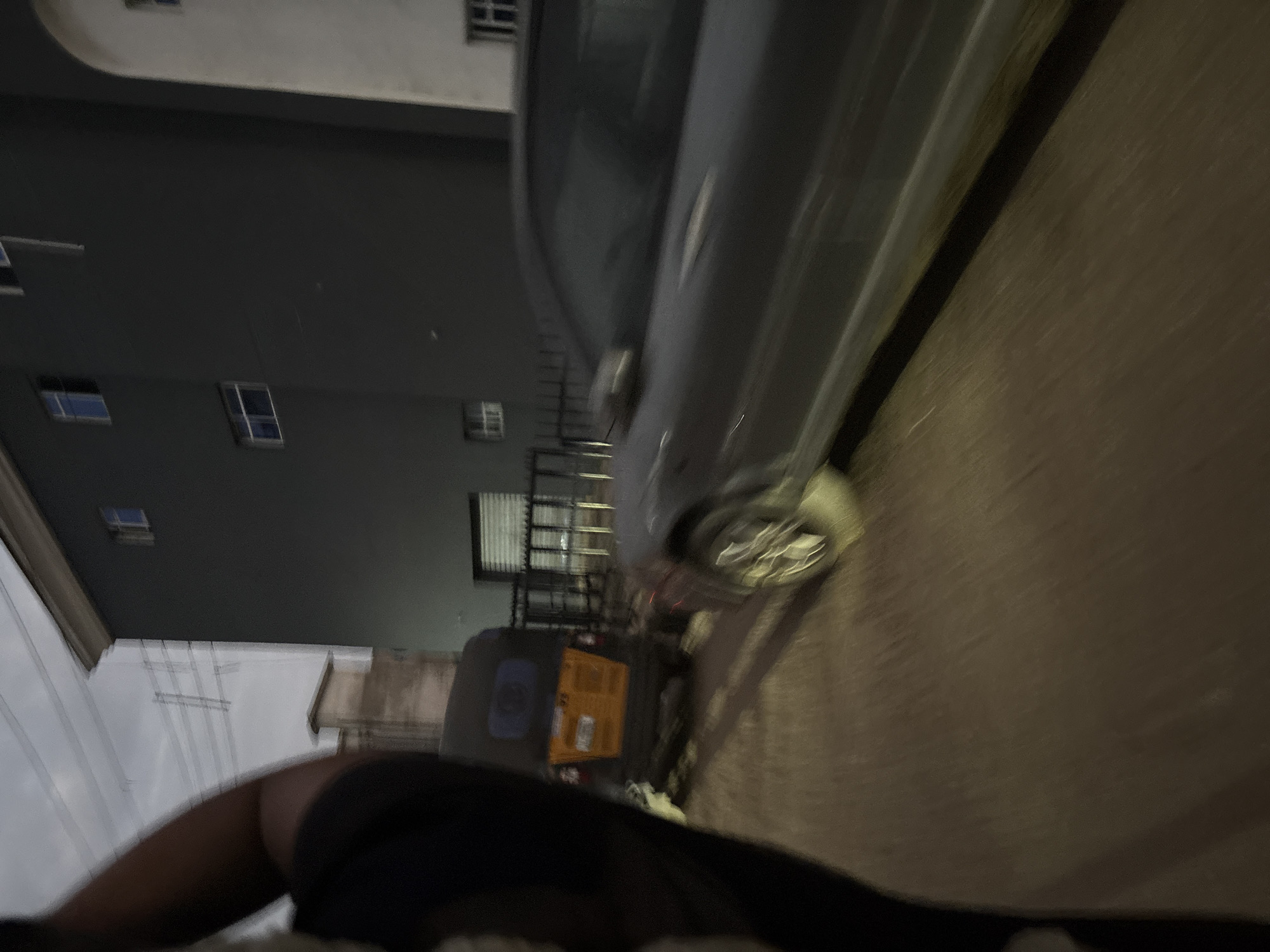 The image shows a street scene in Onitsha, Nigeria. In the foreground, a blurred person's arm and shoulder are visible, indicating the photographer's perspective. To the left, a three-wheeled vehicle, commonly known as a "keke" or "okada," is parked. The keke has a yellow and orange color scheme and a blue tinted window at the back. Beside it, a dark-colored car is moving, captured with motion blur. The car appears to be a sedan. Behind the vehicles, a modern-looking building with dark gray walls and visible windows stands. The sky above is overcast, suggesting it might be evening or a cloudy day. Power lines and poles are also visible in the background, consistent with an urban setting. The ground appears to be paved, likely a road.