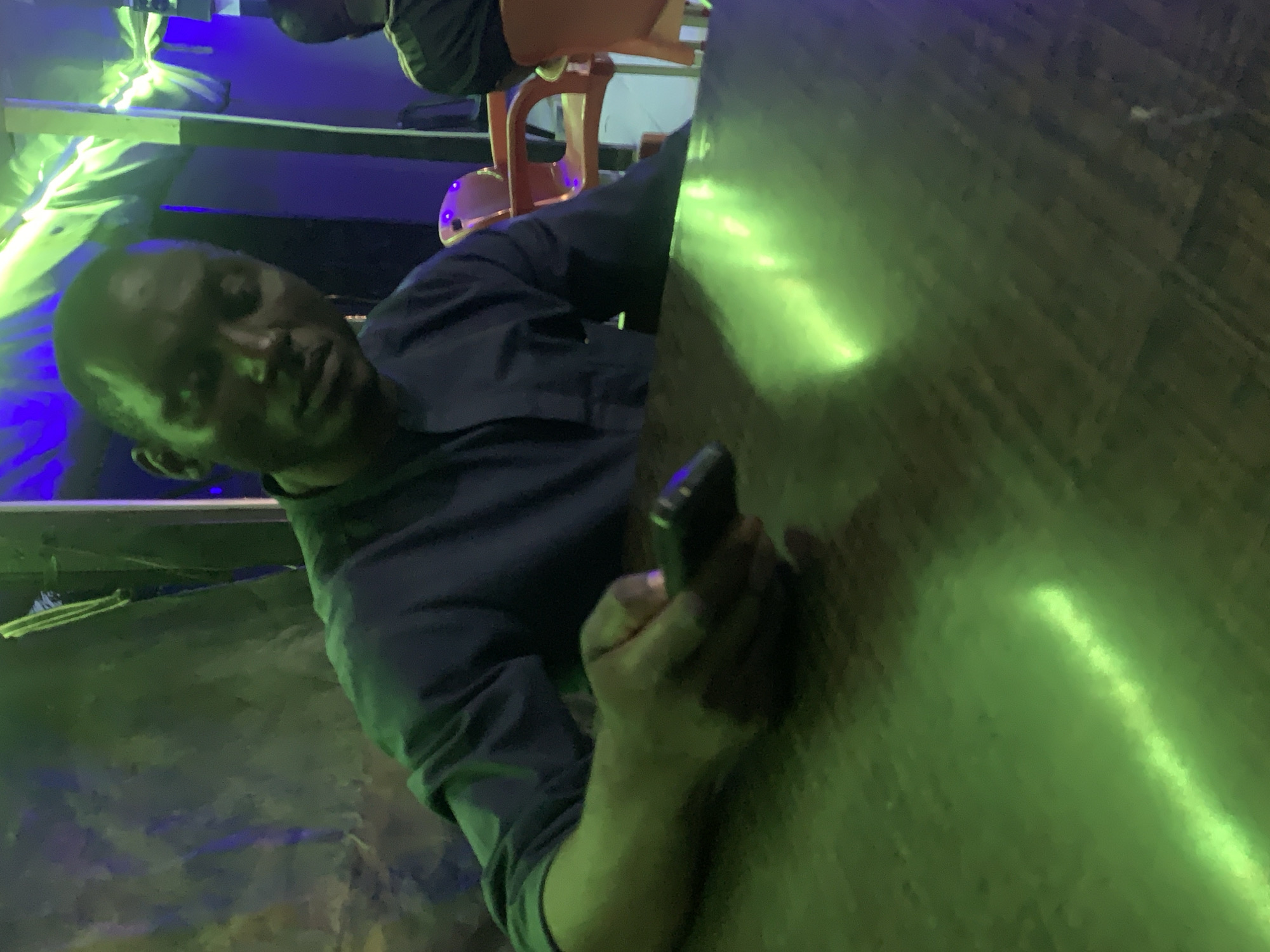 The image features a man captured in a close-up, side-angle view within an indoor setting in Akwanga, Nigeria. The man has dark skin and short dark hair, and he looks directly at the camera with a neutral, possibly slightly tired, expression. He is dressed in a dark-colored long-sleeved shirt or jacket. In his right hand, he holds a dark-colored smartphone.
He appears to be leaning against or sitting adjacent to a dark surface, likely a wooden table or counter, which diagonally bisects the right side of the frame. The environment is dimly lit, predominantly by artificial green and blue lights, suggesting a nighttime scene or an establishment such as a bar, lounge, or club. A distinct linear green light source is visible in the upper left, illuminating the man and elements of the background. Another intense green light reflection is prominent on the table surface. Portions of an orange or brown plastic chair are faintly visible in the blurred background, along with other indistinct dark shapes. The overall atmosphere is characterized by the moody, colored lighting. No discernible text is visible in the image.