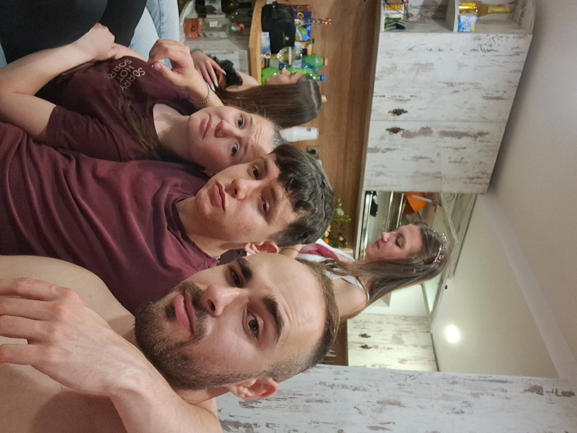 The image shows a group of four individuals indoors, likely in a private residence. The foreground features a man with a beard, looking directly at the camera. Behind him, a young man in a maroon shirt gazes at the lens. Behind him, another young woman in a maroon shirt is looking up at the camera. Further back, a fourth individual is visible, although more indistinctly. The background includes a kitchen counter with assorted items, cabinets, and some kind of decor. There is no clear indication of the exact location in Gabrovo, Bulgaria. There is no clear indication of any actions, interactions or subjects.