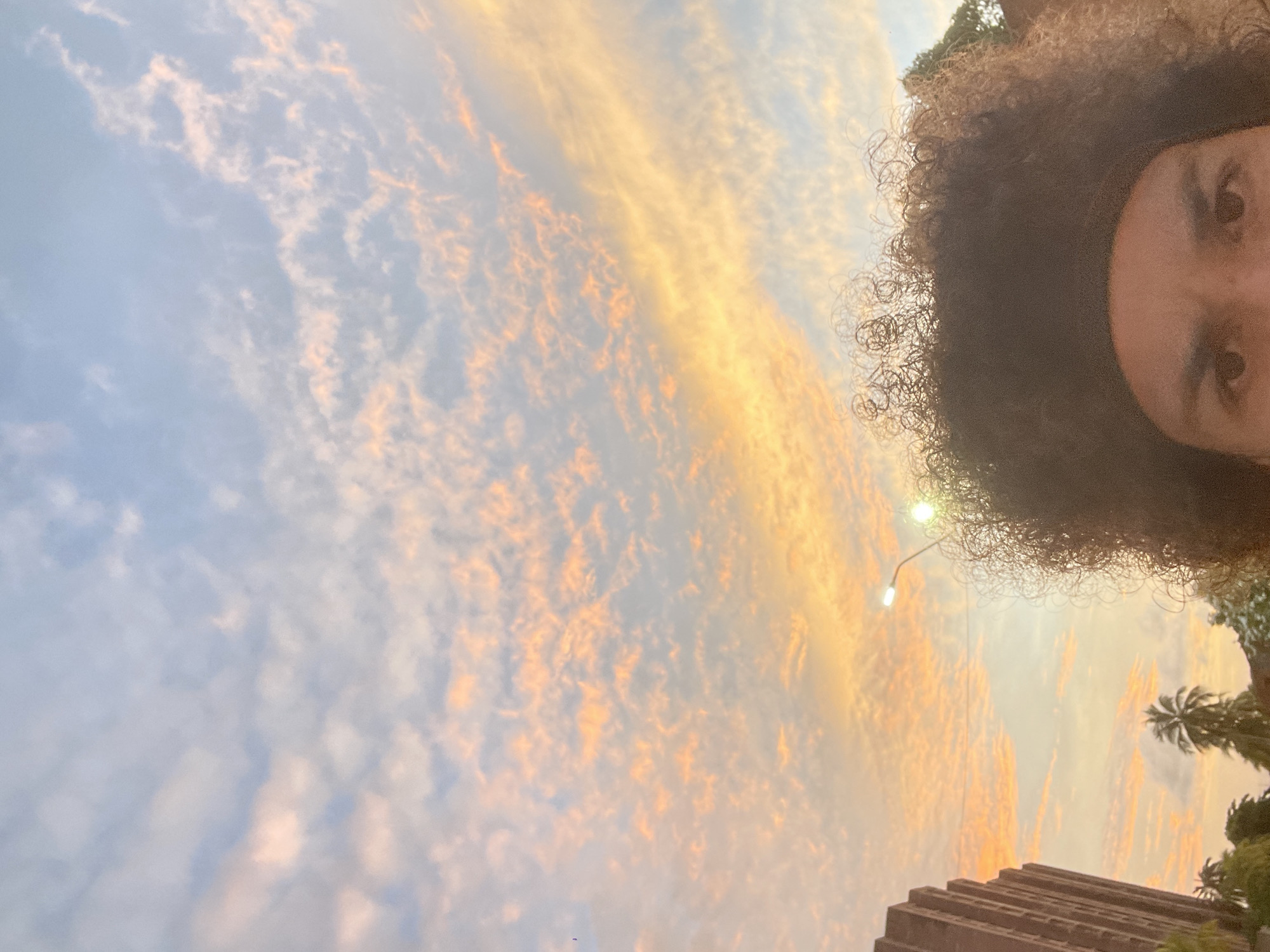 A close-up, low-angle selfie captures the top of a person's head with voluminous, curly, dark brown hair and a dark headband. The person's eyebrows and eyes are visible, looking towards the camera. The background is a vibrant sky at sunset or sunrise, with streaks of pink, orange, and yellow clouds against a light blue sky. To the left, a tall, brown, brick building with many windows is partially visible. In the middle ground, a lamppost with a lit bulb extends upwards. Palm trees and other greenery are visible in the lower portion of the frame. The scene appears to be outdoors in a city, likely Porlamar, Venezuela, given the architecture and tropical vegetation.