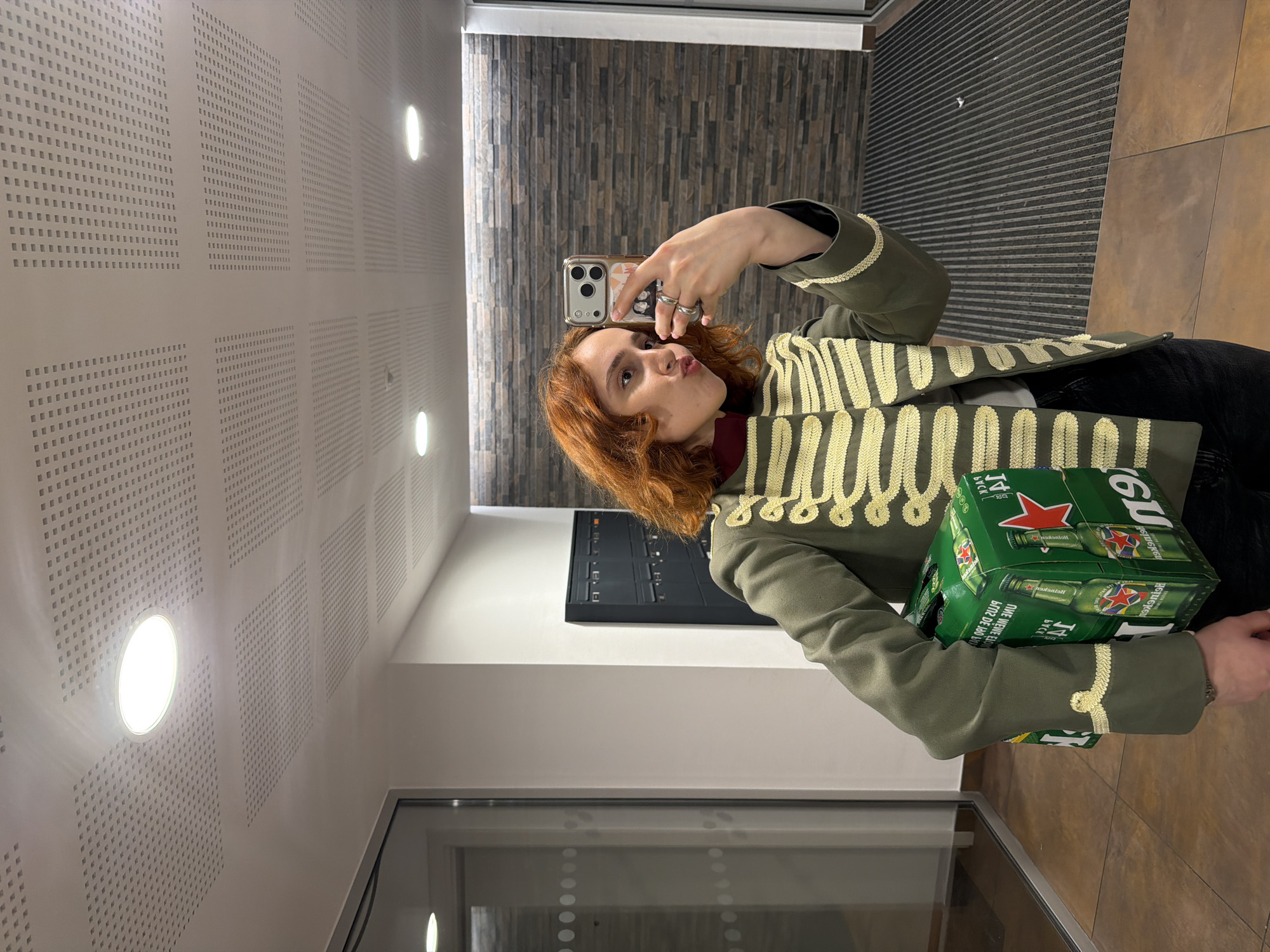 A young woman with auburn hair is taking a selfie in a well-lit, indoor hallway. She is wearing a military-style green jacket with gold trim and a dark turtleneck. In her left arm, she is carrying a six-pack of Heineken beer. Her right hand is holding a smartphone, which is angled to capture her reflection. The hallway has a tiled floor, a drop ceiling with recessed lighting, and a stone-patterned wall in the background. To the left, there is a glass door with round white dots. To the right, there are dark-colored mailboxes. The scene appears to be in an apartment building or office building. The image does not contain any elements that would allow for its specific location to be identified as Strasbourg, France.