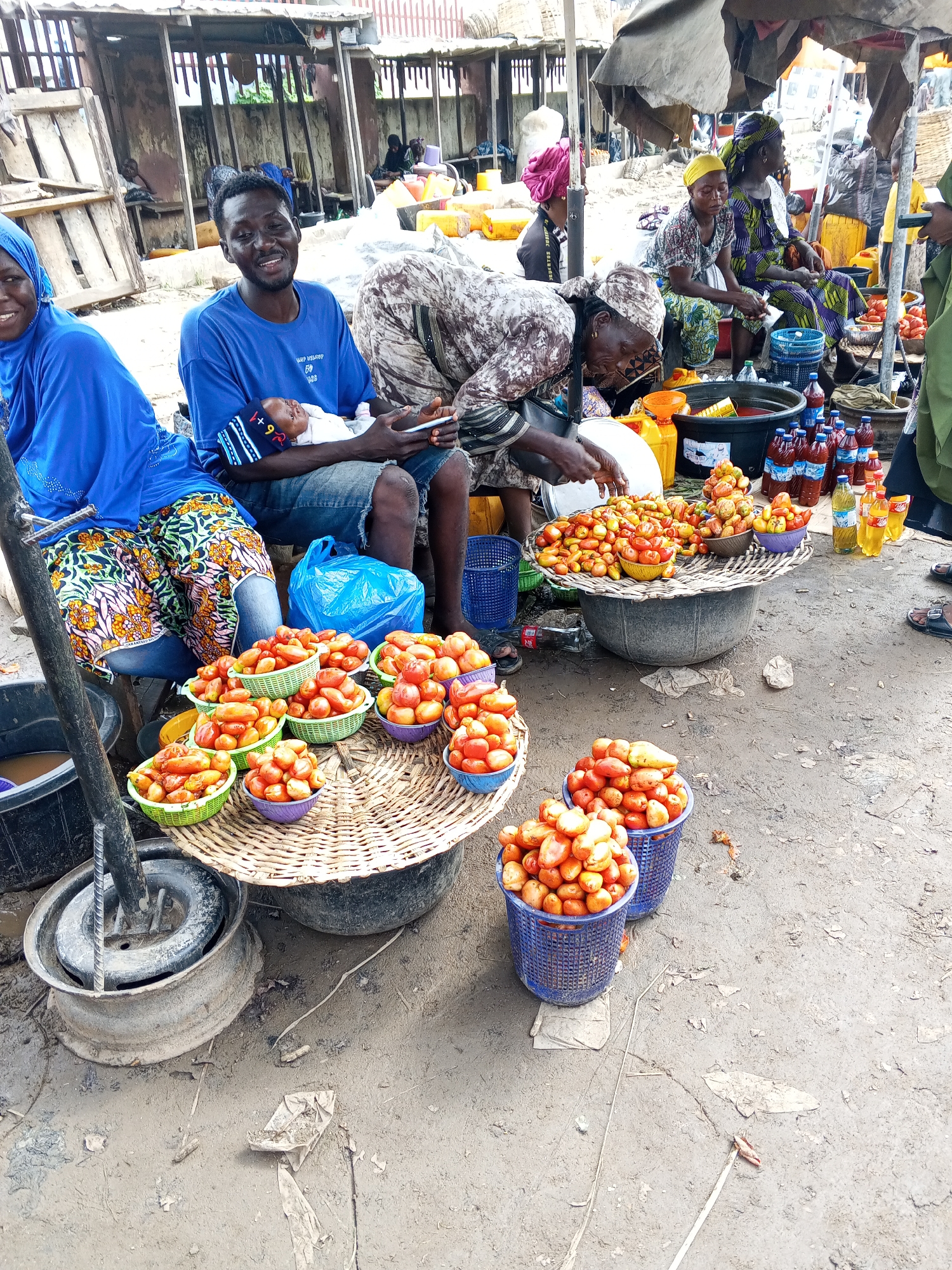 In Lokoja, Nigeria, a busy outdoor market scene unfolds. A man in a blue t-shirt and denim shorts holds a newborn baby wrapped in a striped blanket. Beside him, a woman in a blue hijab and patterned skirt sits, looking towards the viewer with a smile. Several stalls display an abundance of fresh tomatoes in woven baskets of various sizes and colors, including green, purple, and blue. Behind the tomato stalls, bottles of drinks, both clear and reddish-brown, are neatly arranged. A large black basin, possibly for water, is visible to the left, and a woman in a patterned fabric is seated, tending to the market goods. The ground is made of rough, damp earth, with scattered debris. The background reveals more market stalls with wooden structures and covered awnings, suggesting a bustling commercial environment.