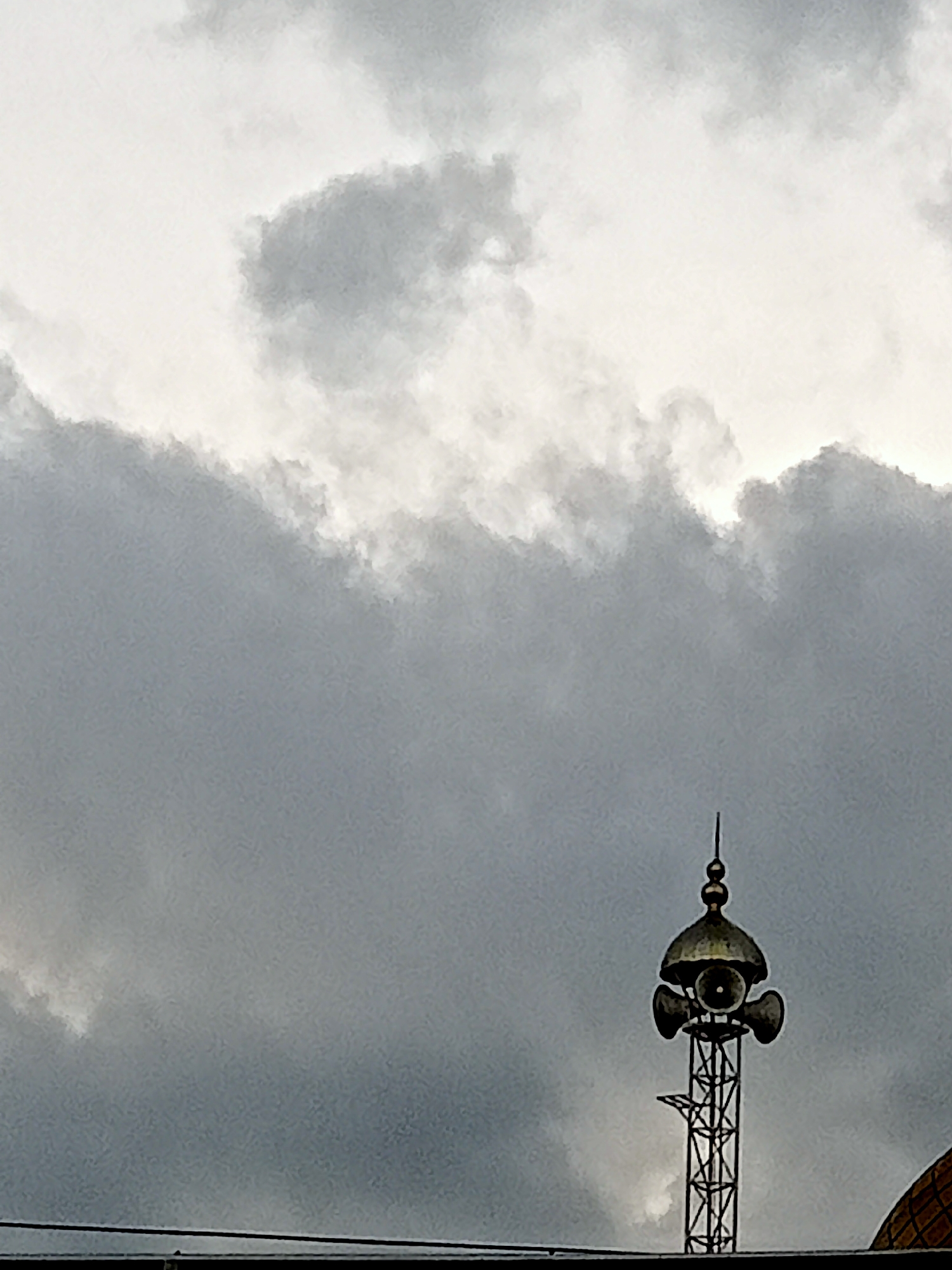 The image shows a telecommunications tower with speakers on top, likely used for broadcasting the adhan (call to prayer) or public announcements. The tower is located in Simo, Indonesia, a town known for its mosques and Islamic heritage. The sky is overcast, with dramatic, dark clouds suggesting an impending storm or heavy rain. A portion of a domed building, possibly a mosque, is visible to the right of the tower. The lower part of the image shows a dark, horizontal line, which could be the roof of a building or a power line. The overall scene is somber and atmospheric, with the tower standing as a sentinel against the elements. There are no people visible in the image.
