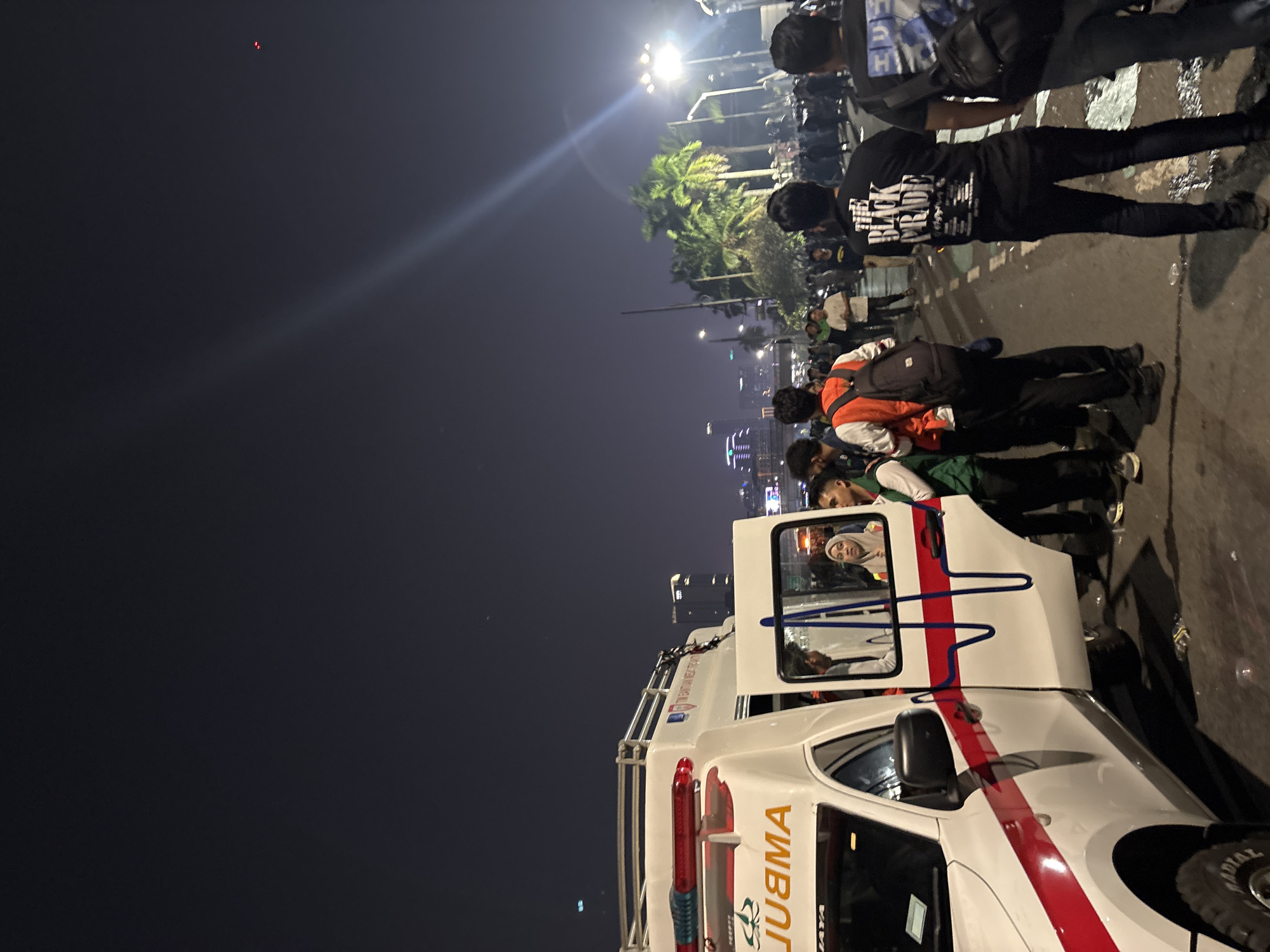 The image captures a nighttime scene on a street, likely in Central Jakarta, Indonesia, with an ambulance prominently featured on the left. The ambulance is white with red and blue accents and text that reads "AMBULANCE" backwards. Several people are gathered around the open doors of the ambulance, some appearing to be medical personnel or bystanders. The background shows a city skyline with illuminated buildings and palm trees, suggesting an urban setting. A crowd of people is visible further down the street, with some wearing backpacks and casual clothing. The lighting suggests it is after dark, with streetlights and building lights illuminating the area. The overall atmosphere appears to be one of activity and potentially an emergency situation given the presence of the ambulance.