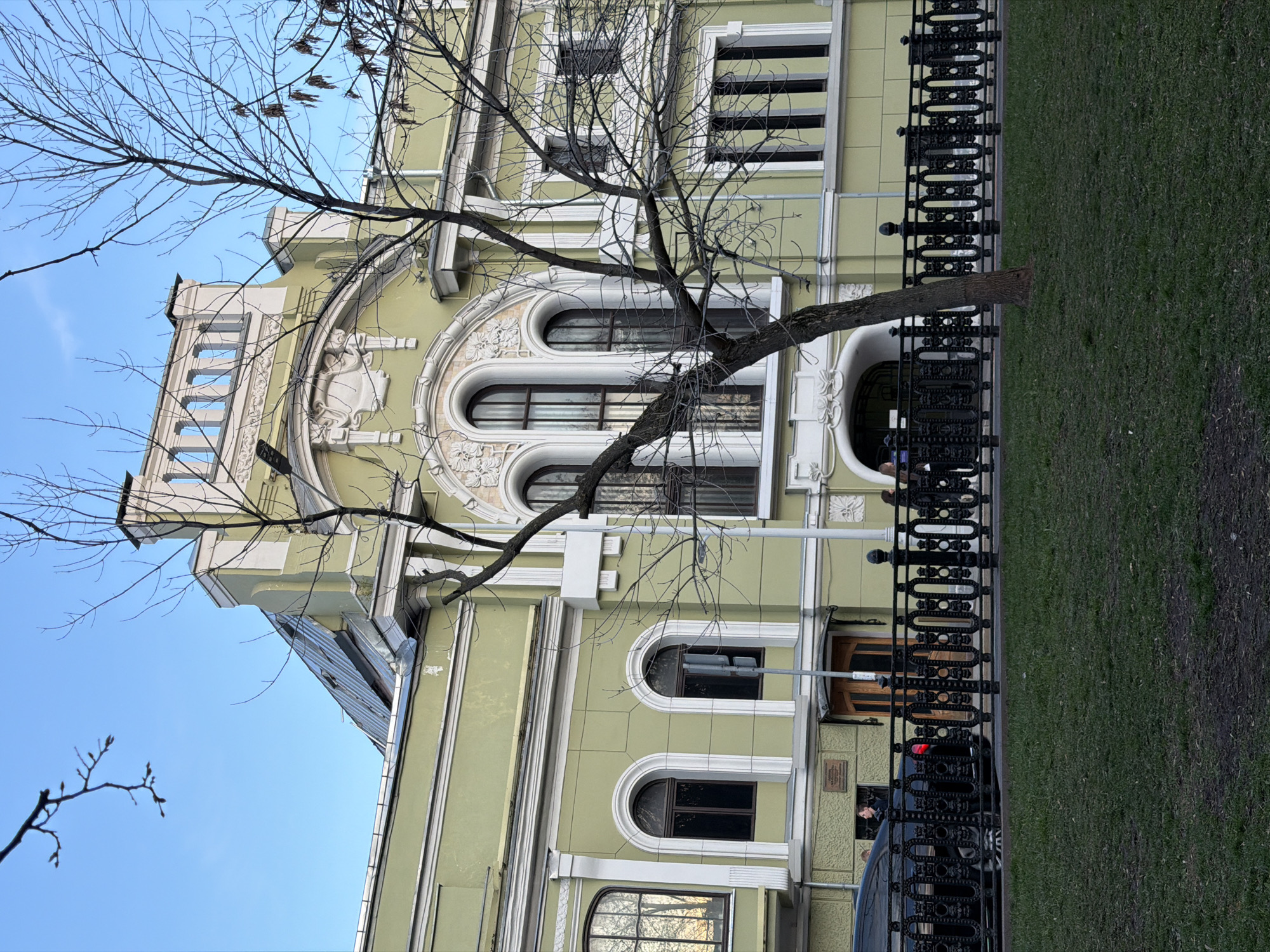 The image depicts a multi-story building in Moscow, Russia. The building is light green with white architectural detailing around the windows and roof. A bare tree is in front of the building. A black wrought iron fence separates the building from a green grassy area. A dark-colored car is parked behind the fence near the building's entrance. The sky is blue. The image appears to be taken from a low angle, looking upwards. The photograph has been rotated 90 degrees counter-clockwise.