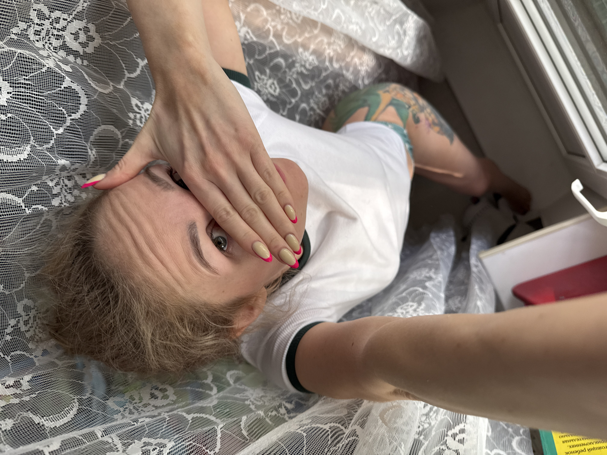 A woman lies on a bed in what appears to be a room with a window. The location is Chaikovsky, Russia. She wears a white t-shirt with dark trim on the collar and sleeves, and her leg is visible with a colorful tattoo. Her face is partially covered by one of her hands, with pink-tipped nails. The bed is covered in white lace fabric. Next to her, near the window, lies a book and a partially visible red mobile phone.