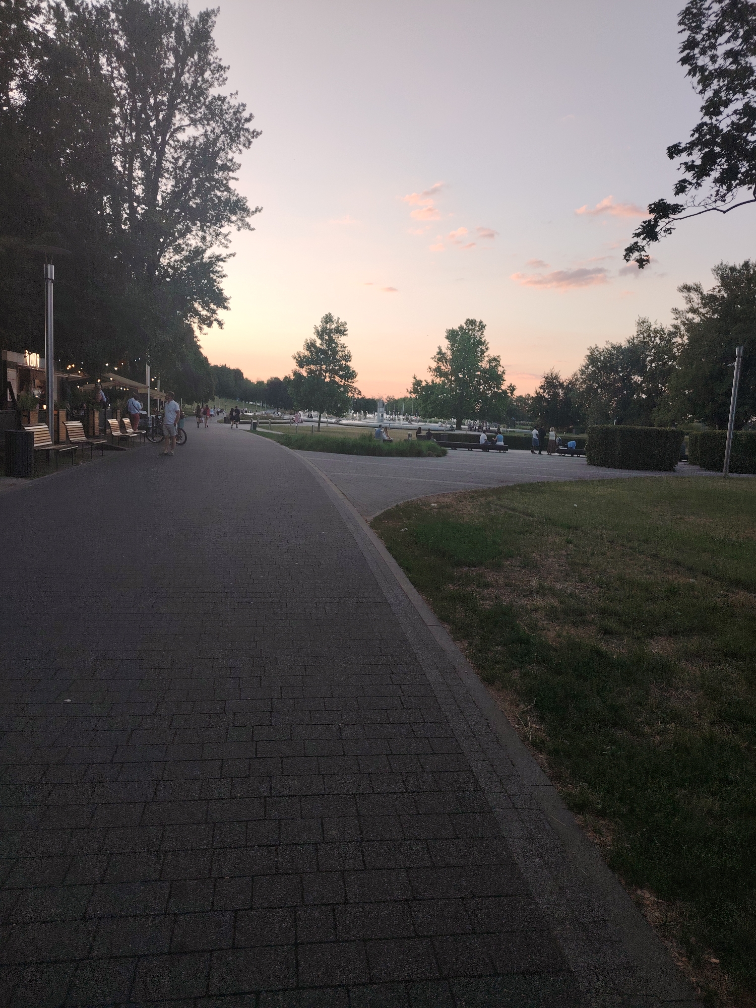 The image depicts a paved pathway in a park-like setting during dusk. The pathway is made of dark gray interlocking bricks and curves to the right, bordered by a strip of concrete and then a grassy area. Several trees with dark green foliage are visible on the left and in the background. A row of wooden benches with metal frames is situated along the left side of the pathway, some of which are occupied by people. Further down the path, more people are walking or standing. In the distance, a fountain is visible, and the sky is a pale gradient of pink and purple, indicating sunset. The overall atmosphere is calm and recreational. This scene is likely located in a public park within Warsaw, Poland, possibly along the Vistula River Boulevards given the urban park setting and open sky.