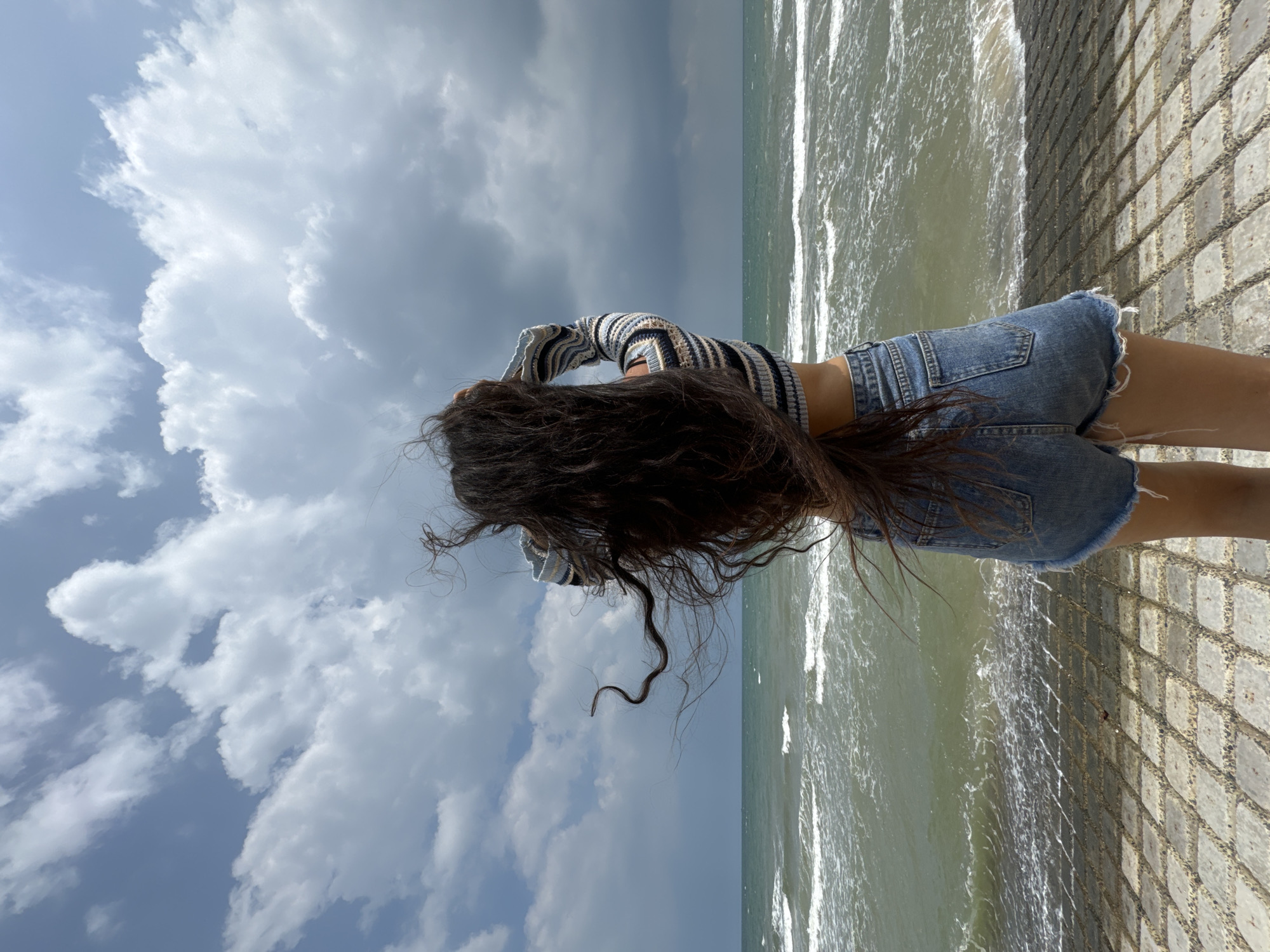 A person stands facing away from the camera, looking out at the sea. Their long, dark hair is windswept. They are wearing a striped crop top and denim shorts. The choppy, greenish-gray sea is in front of them, with white waves breaking near the shore. Behind the person is a paved area made of rectangular stone blocks. The sky above is filled with dramatic, dark gray clouds, suggesting an impending storm, with patches of blue sky visible. The horizon line is clear, separating the sea and sky. The scene appears to be at a coastal location, possibly in Phu Thuy Ward, Vietnam, given the style of paving and the oceanic environment.