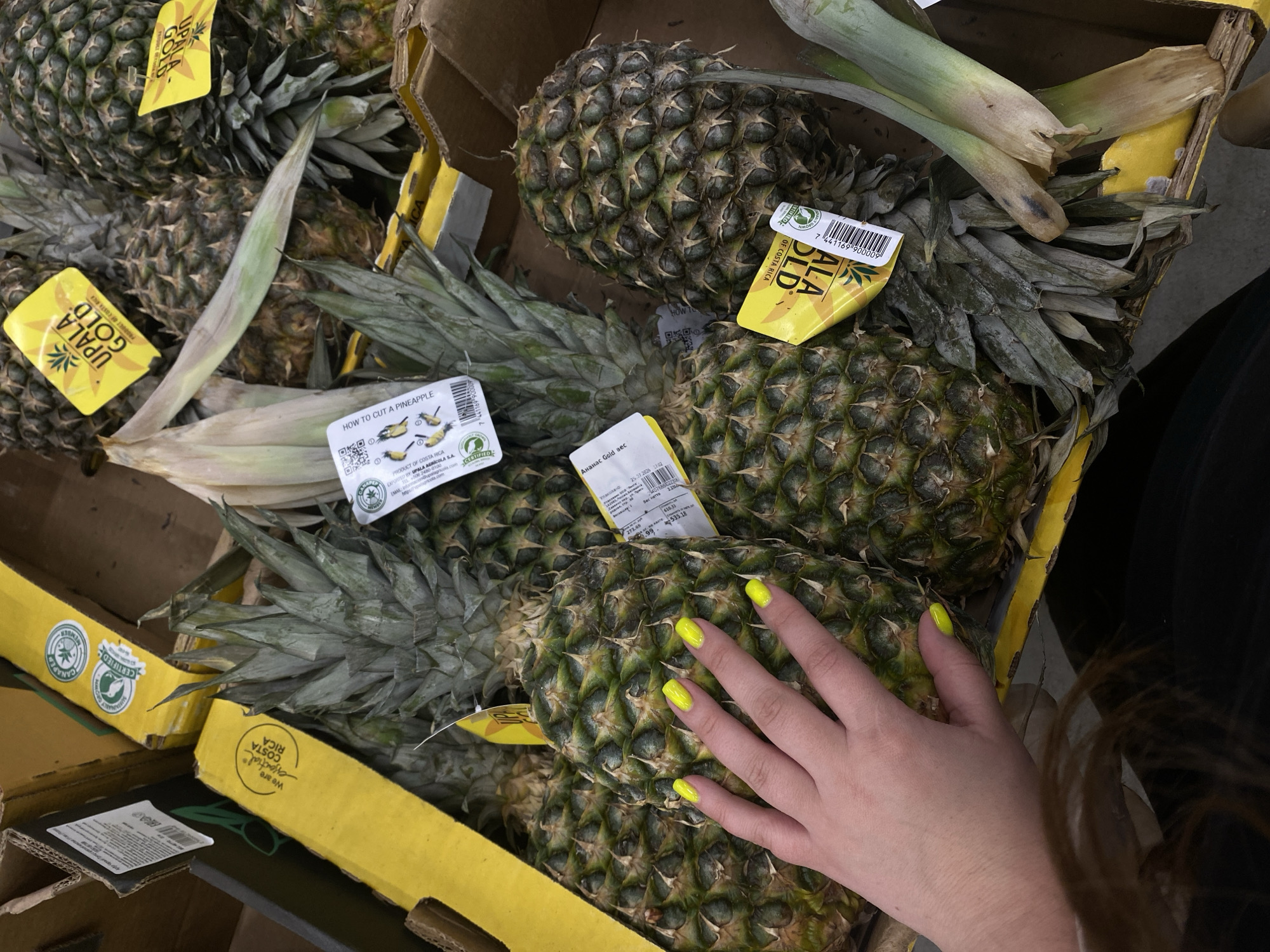 A close-up shot shows a pile of pineapples in cardboard boxes. A person's hand, with bright yellow nail polish, is touching one of the pineapples. Yellow labels with "UPALA GOLD" and "PRODUCT OF COSTA RICA" are visible on some of the fruits. There are also labels with QR codes and instructions on how to cut a pineapple. The pineapples appear fresh, with green leaves at the top and a spiky exterior. The background shows more cardboard boxes, suggesting this is a retail or storage environment. No specific location in Balashikha, Russia, can be determined from the image.