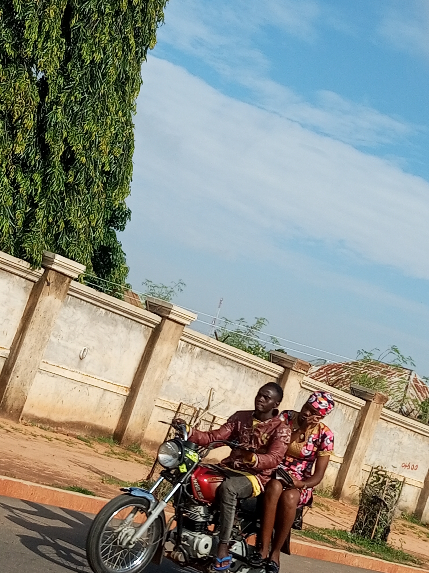 Two people are riding a red motorcycle on a paved road, in the city of Gombe, Nigeria. A man is driving, wearing a maroon, patterned tunic and dark pants. A woman is riding behind him, wearing a colorful, patterned dress and headscarf. The motorcycle is facing left, with the front wheel on the lower left corner of the image. A tall, green tree with dense foliage is on the left side of the image. A light-colored, textured wall with square pillars runs behind the motorcycle. The sky is blue with scattered white clouds.