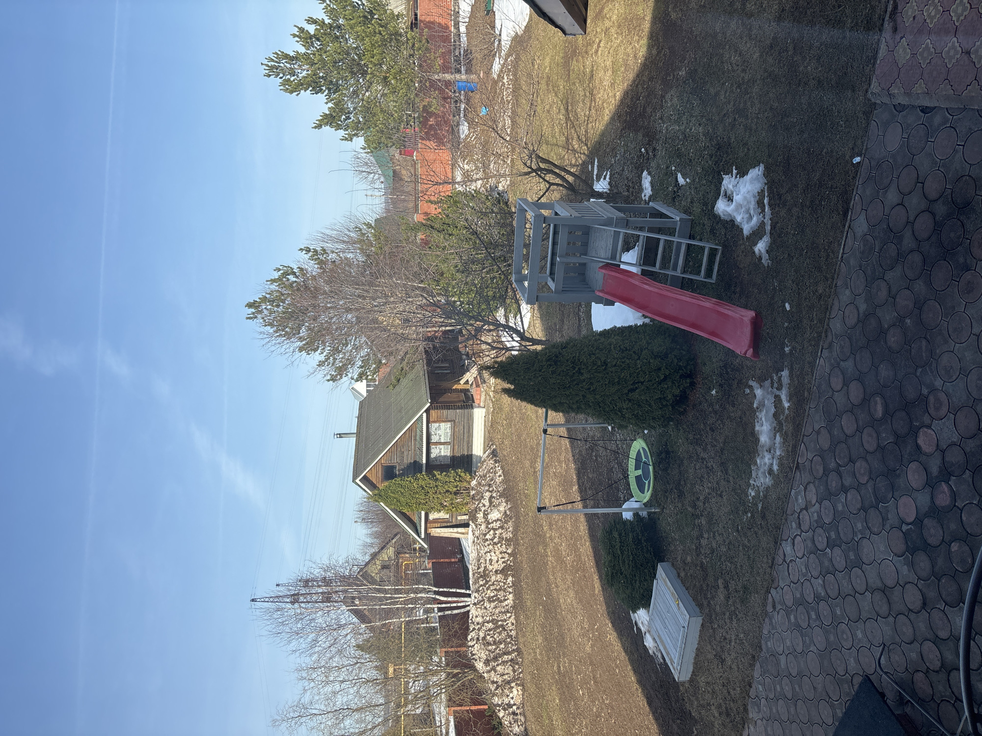 The image shows a backyard scene in Ферма, Russia. In the center of the frame is a playground set with a red slide, a grey wooden climbing structure, and a circular green swing. Patches of melting snow dot the grassy yard. A low stone wall runs along the left side, near a small wooden house with a green roof. Other houses are visible in the distance. The sky is blue with few clouds.