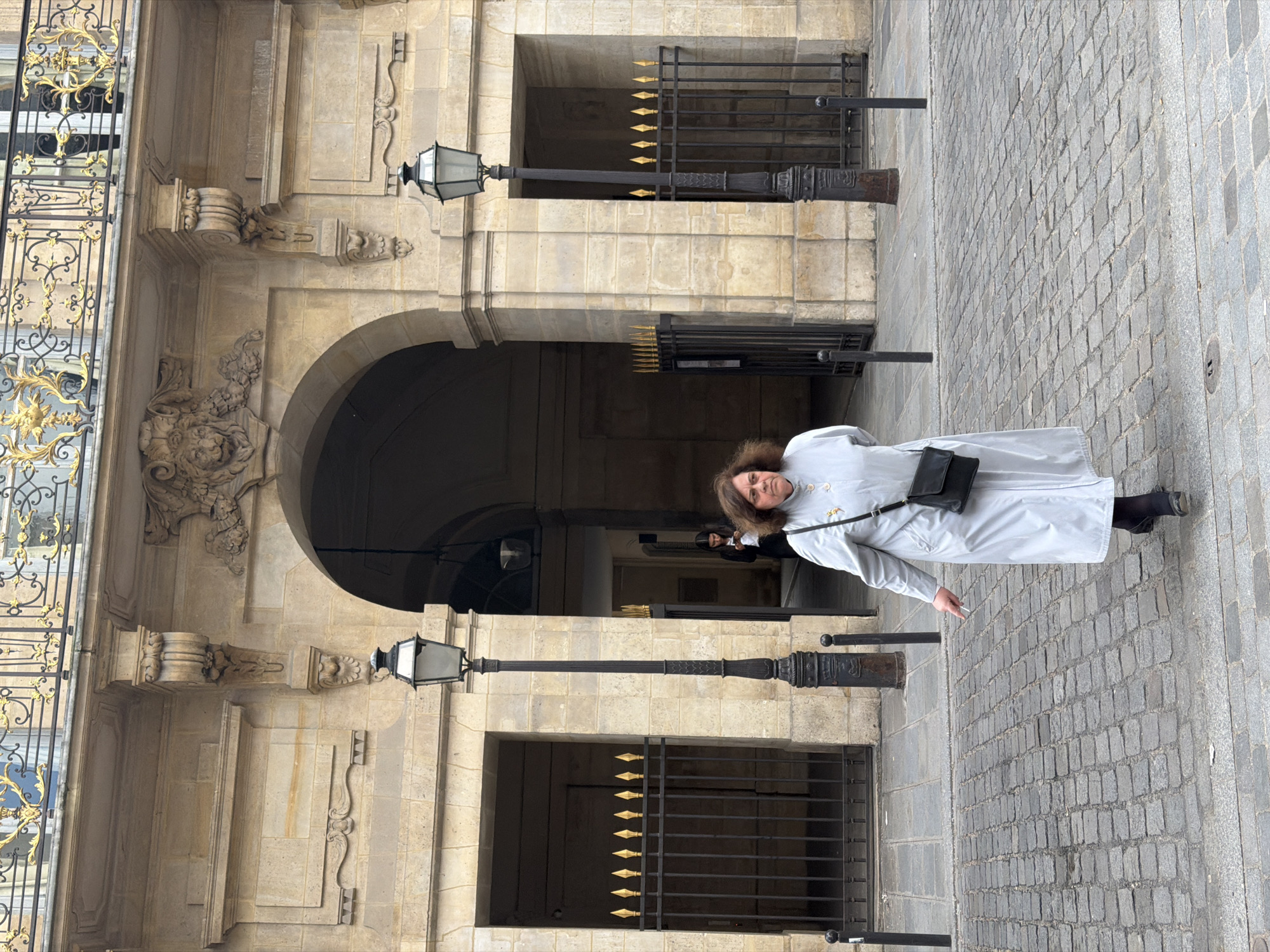 This vertical image depicts a woman standing on a cobblestone street in front of a grand stone building, characteristic of Parisian architecture. The woman, with shoulder-length brown hair, is wearing a light grey or off-white long coat, black trousers, and black boots. A black crossbody bag is slung over her shoulder, and she holds what appears to be a cigarette in her right hand. Her expression is neutral.
The building behind her features light-colored stone blocks, intricate carvings, and decorative elements such as corbels and ornate moldings. It has large arched passages or entryways, with one particularly prominent archway behind the woman. Black wrought iron gates with golden, spear-tipped finials are integrated into the facade, securing openings. Two tall, ornate black street lamps with glass lanterns stand prominently in the foreground, between the woman and the building. To the left, a section of an elaborate golden wrought iron railing or balcony adds a touch of luxury to the architecture. The ground is paved with grey cobblestones laid in a diagonal pattern. The scene appears to be set during daytime, under what seems to be an overcast sky or in a shadowed area, given the soft, diffuse lighting and lack of harsh shadows. There is no visible text in the image.