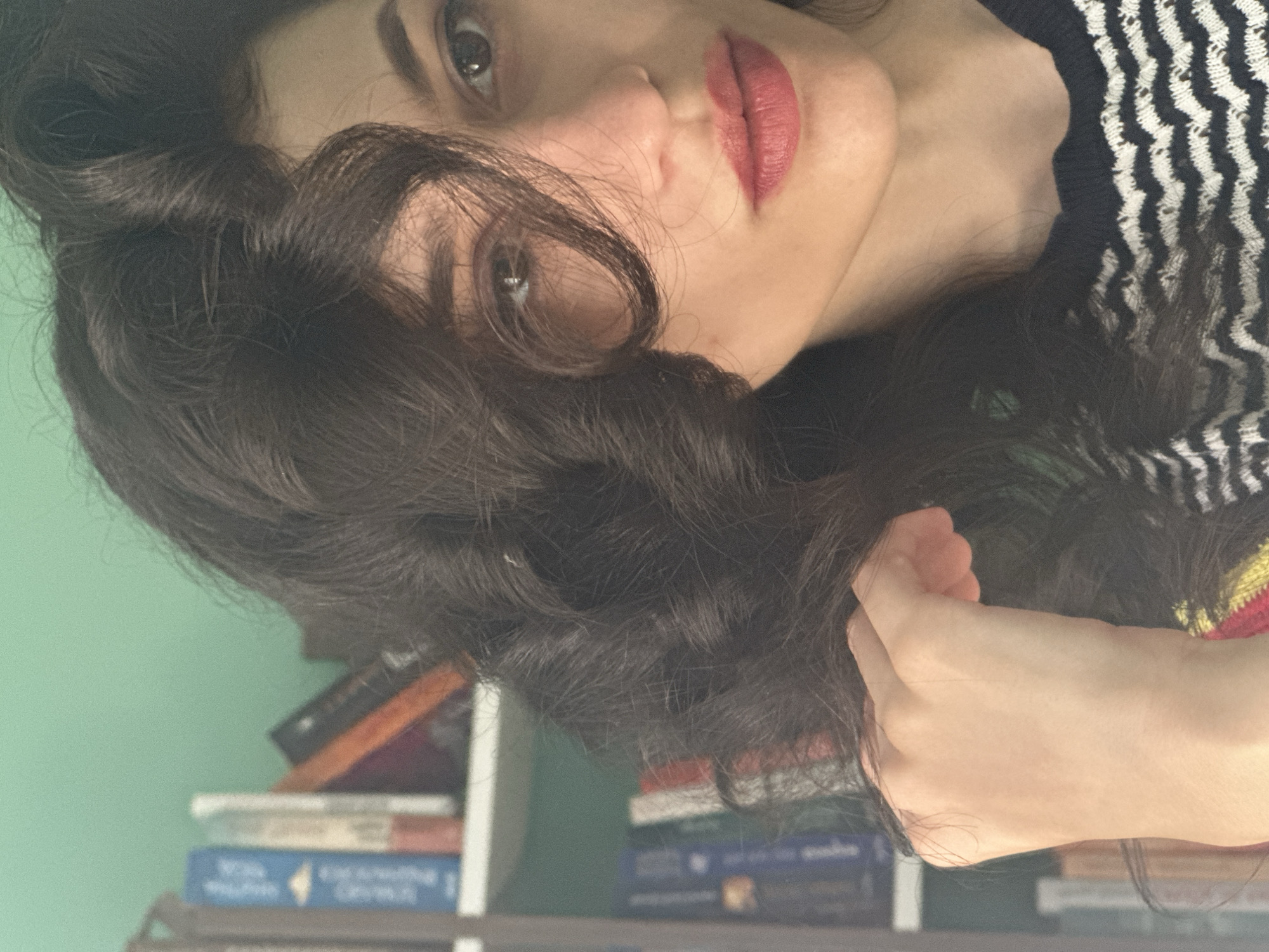A close-up shot captures a woman's face, partially obscured by her dark, wavy hair. She has fair skin, dark eyebrows, and is wearing pink lipstick. Her right eye is visible, looking towards the camera. Her left hand is raised, touching her hair. She is wearing a black and white striped sweater. In the background, a bookshelf filled with books is visible against a green wall. The overall lighting suggests an indoor setting. There is no visible indication of the location being in Sofia, Bulgaria.