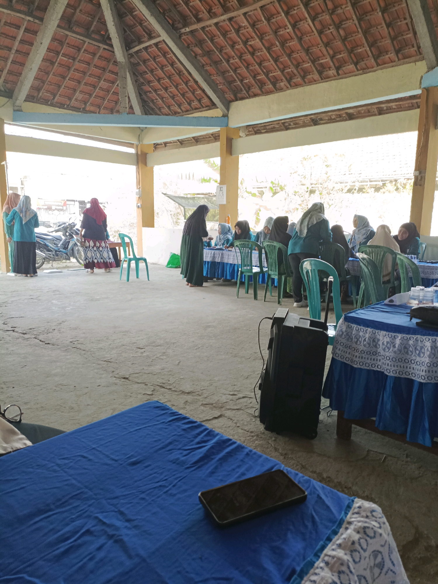 The image depicts an outdoor gathering or event taking place under a covered structure with a tiled roof, possibly in Rancang Kencono, Indonesia. The ground is paved concrete. In the foreground, a table covered with a blue cloth is visible, with a smartphone and a pair of glasses on it. Further back, another table draped in a blue cloth with a decorative white trim is set up. A black speaker unit stands on the concrete floor.
Several individuals, predominantly women wearing hijabs and modest clothing, are present. Some are seated at tables, while others are standing and interacting. One woman in a black abaya and hijab is standing near a table on the right. To the left, three women are standing near a parked motorcycle. The overall atmosphere appears to be that of a community event or meeting. The background shows some foliage and buildings, suggesting a semi-urban or rural setting.