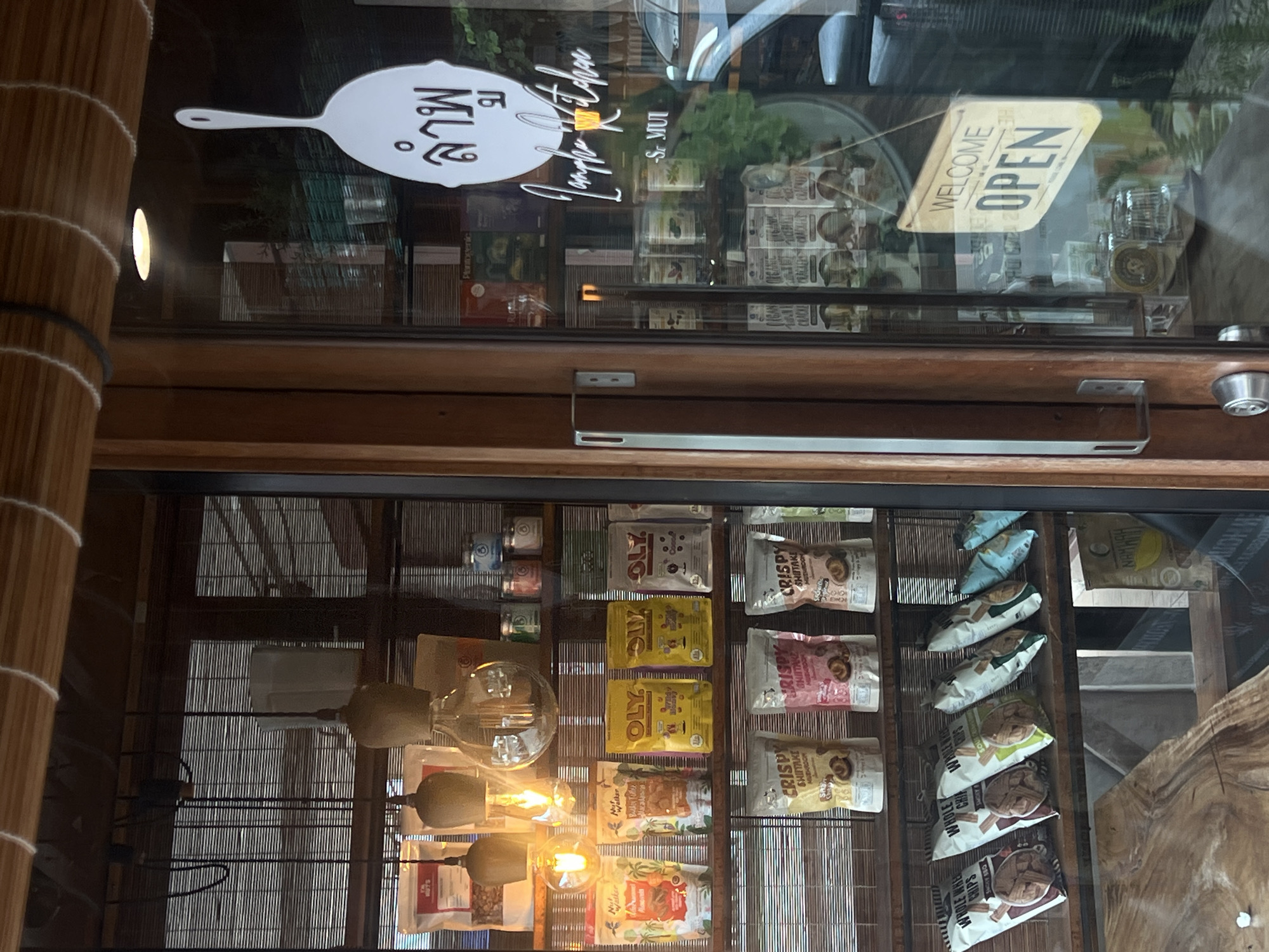 The image captures the interior of a shop or cafe, viewed through a glass window or door. The scene is dominated by shelves neatly stocked with various packaged food products. Prominently displayed are pouches labeled "OLY," "CRISPY SHIITAKE MUSHROOM," and "WHOLE WHEAT CHIPS." Other visible product labels include "Plantogenic" and partially obscured text like "NAMHAH KOWOL SMOKY."
A decorative light fixture with several exposed filament bulbs hangs in the foreground, casting a warm glow. Affixed to the glass is a distinctive white sign shaped like a spatula or frying pan, featuring Thai script "ลำพู" (Lamphu), "Lamphu Kitchen" in a stylized cursive font, and "-S MUi," which likely references Koh Samui. Below this, a rectangular sign hangs, clearly stating "WELCOME OPEN." The interior decor includes wooden elements and a textured wall resembling bamboo or wicker. No people are visible in the image. The lighting suggests it is daytime, with artificial lights supplementing the natural light. The setting implies a specialty food store or a cafe offering local products.
