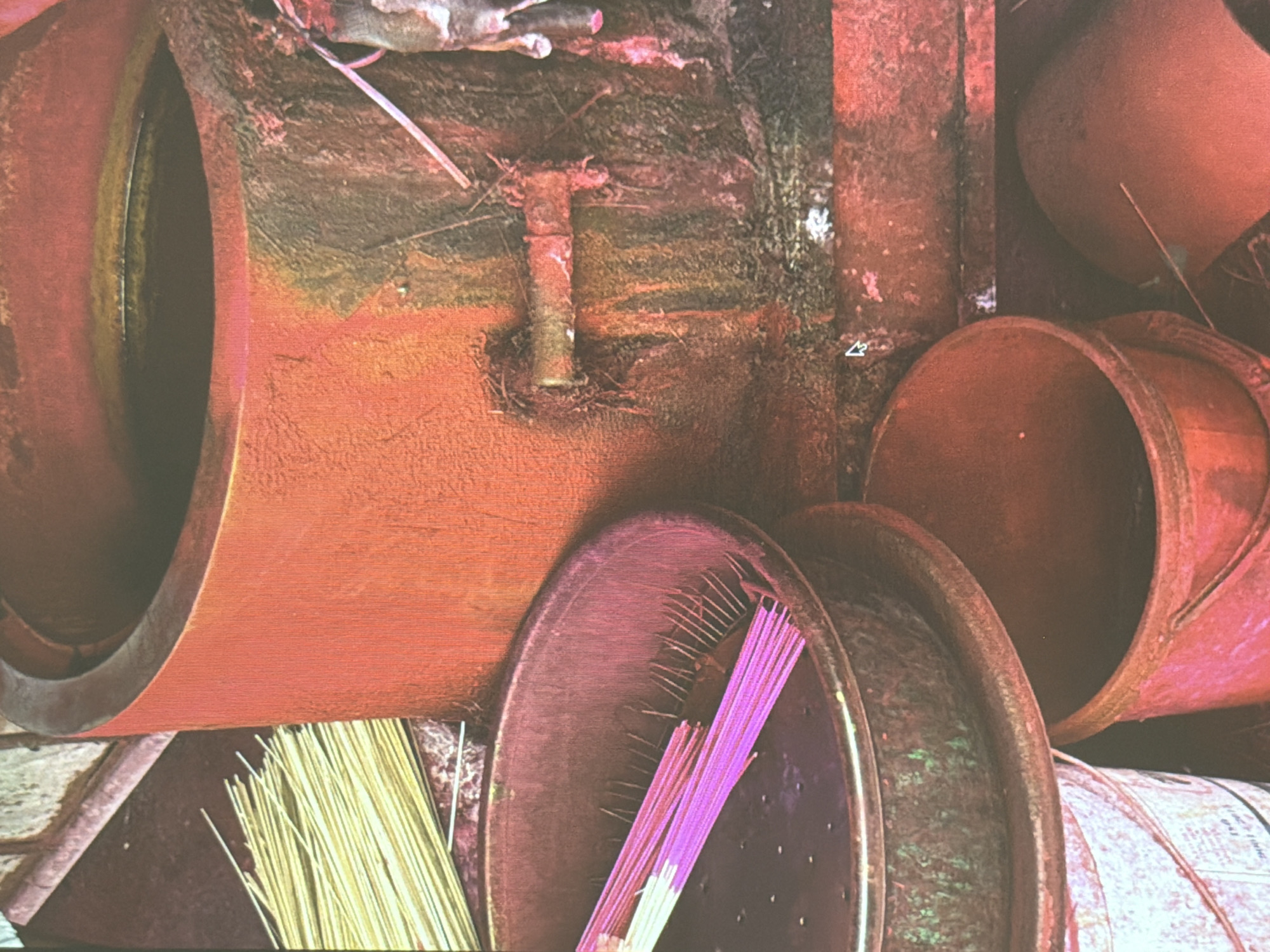 The image shows an arrangement of cylindrical objects and incense in Đà Nẵng, Vietnam. Dominating the composition are several large, reddish-brown cylindrical structures, possibly industrial pipes or large pots, positioned at various angles. One large cylinder occupies the left side, its circular opening facing the viewer. To the right and below, are other cylinders, some stacked and overlapping.
At the bottom center, two circular trays or baskets are visible. One contains yellow incense sticks, and the other, sticks that are purple. The sticks are arranged in bundles. The overall scene appears to be cluttered and possibly industrial or storage-related, potentially outdoors, evidenced by the slightly weathered and aged appearance of the objects. A T-shaped metal joint or pipe segment is also visible.