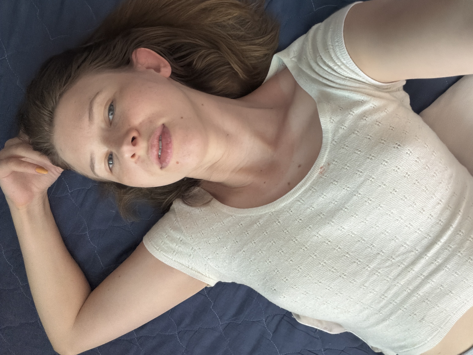 This is a close-up, overhead shot of a young woman lying on her back on a dark blue quilted surface, likely a bed or couch. She has light brown hair that is spread out around her head. She is wearing a cream-colored, short-sleeved top with a textured pattern. Her left arm is bent at the elbow, with her hand resting near her head. Her right arm is partially visible, also bent. The woman is looking upwards and slightly towards the camera, with a neutral expression. Her face is well-lit, revealing some freckles. The background is a dark blue quilted fabric. There are no visible objects, text, or indications of the time of day, weather, or specific location beyond the provided context.