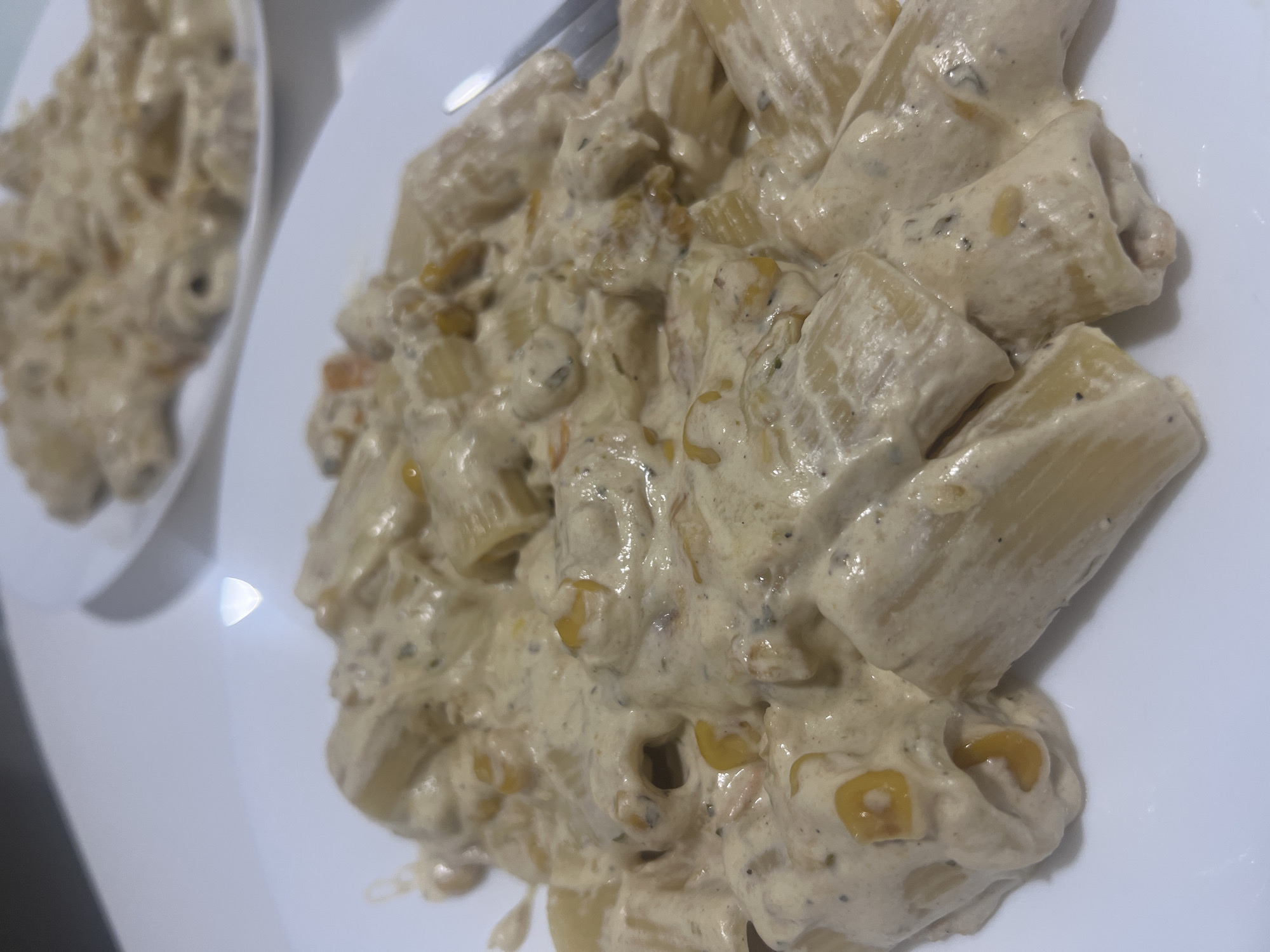 The image shows two white plates of rigatoni pasta covered in a creamy sauce with what appear to be corn kernels. The pasta dishes are presented in an indoor setting. There are no people visible in the frame. The environment is plain and suggests a domestic or restaurant setting. There is no indication of the location being Mogán, Spain.