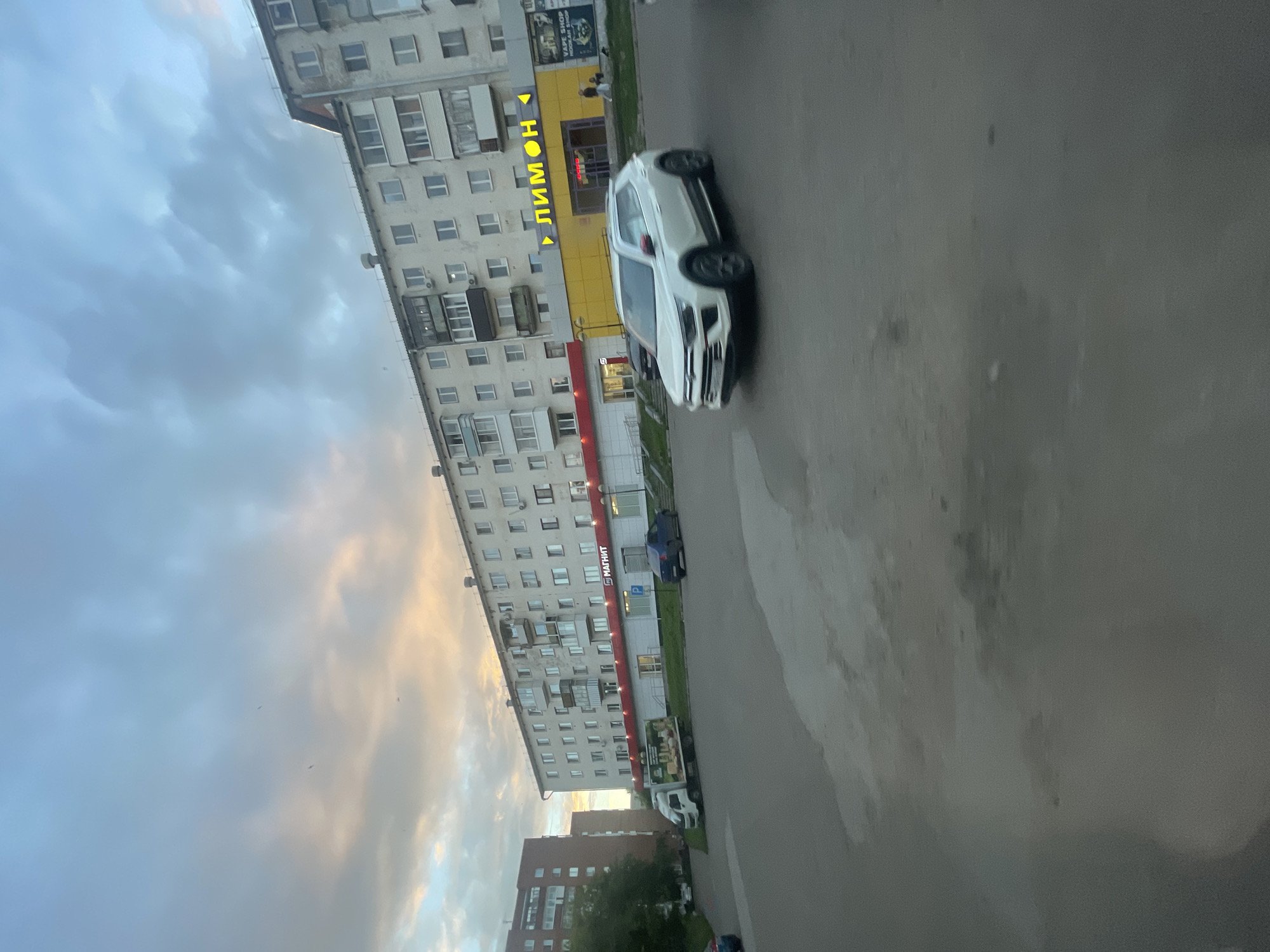 The image depicts a street scene in Votkinsk, Russia, during what appears to be late afternoon or early evening, with the sky showing a mix of gray clouds and warm, golden hues from the setting sun. The primary subjects are a white car, likely a crossover SUV, parked on the right side of the frame, and a long, multi-story residential building that dominates the left side.
The building is typical of Soviet-era architecture, with a light-colored facade, numerous windows, and balconies. Several storefronts are visible at the ground level of the building. One prominent sign, written in yellow Cyrillic letters, says "ЛИМОН" (LIMON), suggesting a store or establishment named "Lemon." Another sign, in red Cyrillic, reads "МАГНИТ" (MAGNET), which is a well-known Russian supermarket chain. A blue car is parked in front of the MAGNET store. Further down the street, a white truck with a large advertisement on its side is visible.
The overall atmosphere of the scene is quiet and ordinary, capturing a moment of everyday life in a residential urban area. The lighting suggests a time when the day is winding down, casting long shadows and creating a soft glow. There are no people clearly visible in a way that suggests specific activity, other than perhaps a few figures in the distance near the storefronts, whose presence is indistinct. The weather appears to be partly cloudy, with the sunlight breaking through the clouds. The image is oriented vertically.