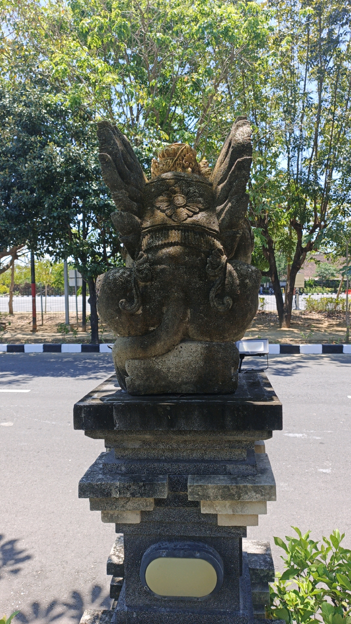 A stone statue of a mythical creature, possibly a garuda, stands atop a carved stone pedestal in what appears to be a public area in Denpasar, Indonesia. The statue is dark gray and weathered, with prominent wings, a detailed headdress, and a decorative element on its chest resembling a flower. It is positioned outdoors, with lush green trees and blue sky visible in the background. The pedestal is made of multiple tiers of carved stone, with a dark, rectangular base. A paved road with white lines and a black and white striped curb is in the foreground, suggesting it is located near a street or pathway. Sunlight casts shadows of foliage onto the road and the lower part of the pedestal. The overall impression is that of a cultural or religious artifact in a tropical urban environment.