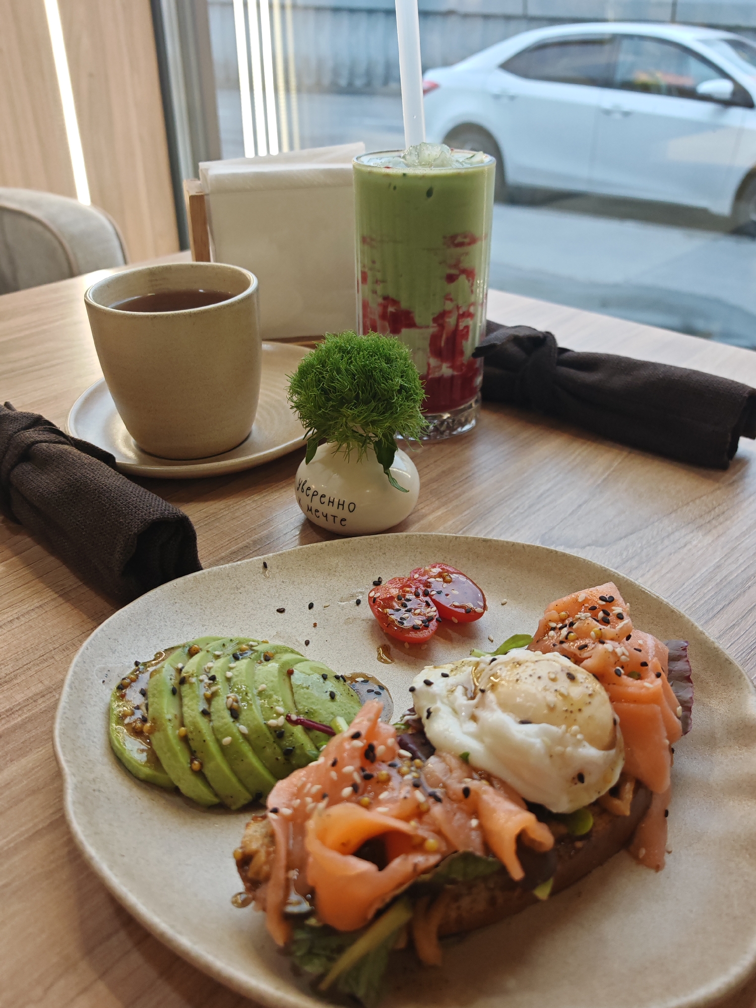 A close-up shot shows a meal on a wooden table. In the foreground, a light-colored ceramic plate holds a dish consisting of a piece of toast topped with lettuce, smoked salmon, and a poached egg, sprinkled with sesame seeds. To the left of the toast, sliced avocado with sesame seeds and a drizzle of sauce is arranged. Two halved cherry tomatoes with sesame seeds and sauce are placed above the toast. In the background, a ceramic mug of dark tea sits on a saucer. To the right of the tea is a tall glass filled with a green beverage layered with red syrup and ice, with a straw. A small decorative white vase with a green fluffy plant and text in Cyrillic script sits between the tea and the beverage. A dark cloth napkin is tied around a rolled napkin to the left of the plate, and another similar napkin is tied around a rolled napkin behind the glass. Through a window in the background, a white car is visible on a street, suggesting an urban setting, possibly in Novy Urengoy, Russia, given the context of a restaurant or cafe. The lighting suggests it is daytime.