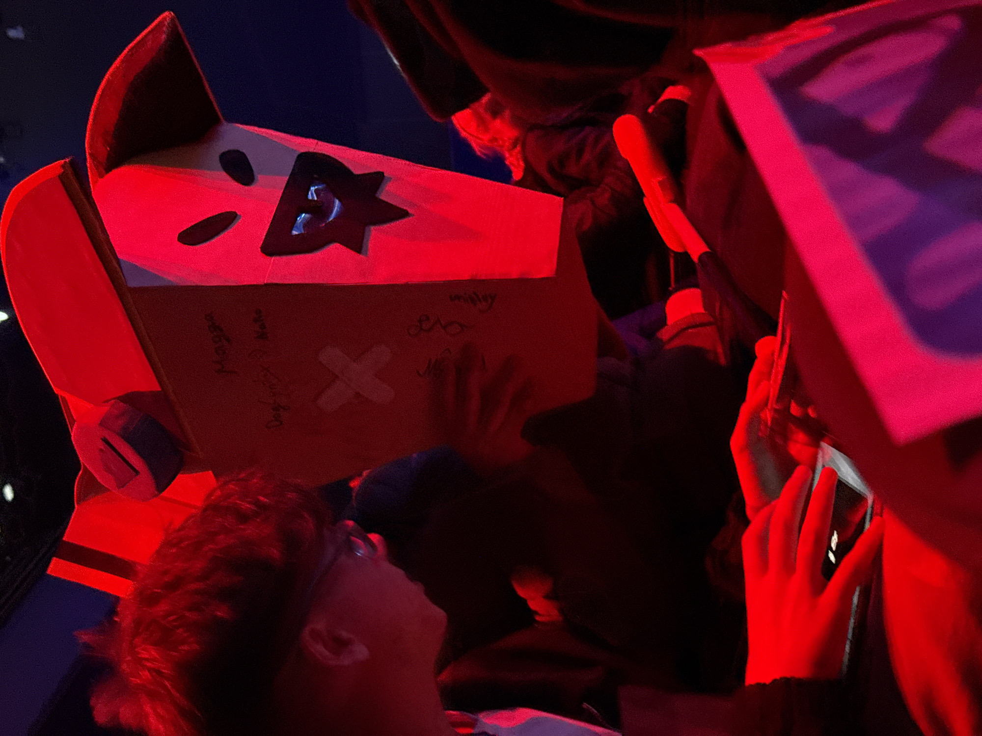 A person wearing glasses and a white shirt is seen looking upwards at a cardboard box crafted into the shape of a robot head, adorned with cut-out shapes resembling eyes and a mouth. The box is signed with names and includes a drawing of a cross. A second person, whose hand is visible, is reaching out to touch the box. A third person is seen using a cell phone to take a picture of the box. The scene is illuminated by bright red lighting. While the location in Berlin, Germany, cannot be definitively confirmed from the provided visual information, the scene suggests an event or gathering where interactive elements like a signed and crafted box are featured.