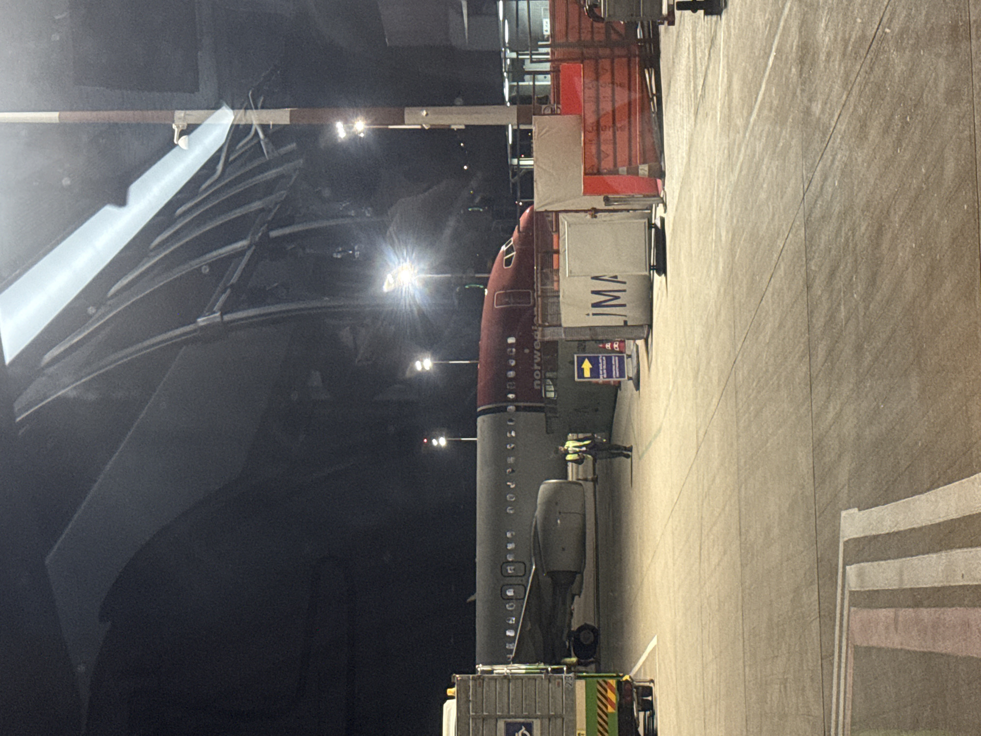 The image shows a Norwegian aircraft parked at an airport gate at night. The aircraft is connected to a jet bridge. Ground crew personnel are visible near the aircraft. The airport structure features a modern, curved roof design. "LIMA" is visible on the side of the jet bridge.