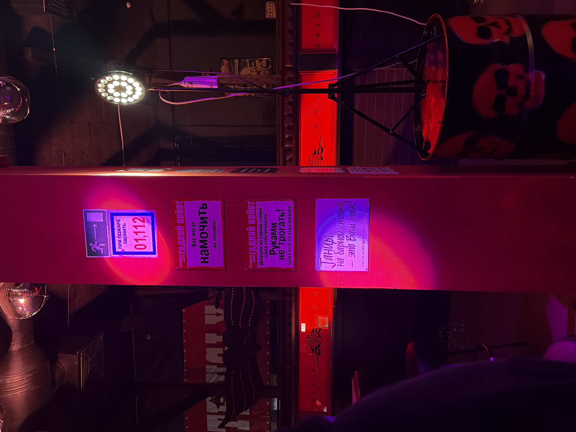 This media file captures an indoor scene, likely a bar or club in Kazan, Russia, characterized by its dim, atmospheric lighting. The space is illuminated primarily by red and purple light sources, casting a vibrant glow, particularly on a central red vertical pillar.
The scene features various objects contributing to a lively, somewhat edgy ambiance. A circular LED light fixture is prominently visible at the top, along with what appears to be part of an industrial ventilation system on the left. On the right, a black barrel decorated with red skulls rests on a stand, reinforcing a grunge or alternative aesthetic. Background elements include dark walls and structural beams. The text "COYOTE UGLY" is visible on two vertically oriented red panels, strongly suggesting the location is a Coyote Ugly Saloon.
No people are directly visible, but the presence of bar rules and warnings implies an active environment where patrons and staff would typically be present.
Notable details include the time of day, which appears to be night, indicated by the artificial lighting. The overall mood conveyed is energetic and possibly rebellious, consistent with the décor and visible rules.
Visible text, primarily in Russian, includes:
* On a blue sign: "ПРИ ПОЖАРЕ ЗВОНИТЬ 01,112" which translates to "In case of fire call 01,112".
* On a pink sign: "ПРАВДА ГАДКИЙ КОЙОТ" (Truth/Rules of the Ugly Coyote) and "Вас могут намочить (не случайно)" (You might get wet (not by accident)).
* On another pink sign: "ПРАВДА ГАДКИЙ КОЙОТ" (Truth/Rules of the Ugly Coyote), "Девушки на барной стойке - наша собственность! Руками не трогать! (даже если это ваша девушка)" (Girls on the bar counter are our property! Do not touch with hands! (even if it's your girlfriend)).
* On a purple sign: "Танцы на барной стойке, это Ваш риск! БАР НЕ НЕСЕТ ЗА ЭТО ОТВЕТСТВЕННОСТЬ" (Dancing on the bar counter, this is your risk! THE BAR IS NOT RESPONSIBLE FOR THIS).
* Vertically on red panels: "COYOTE UGLY".