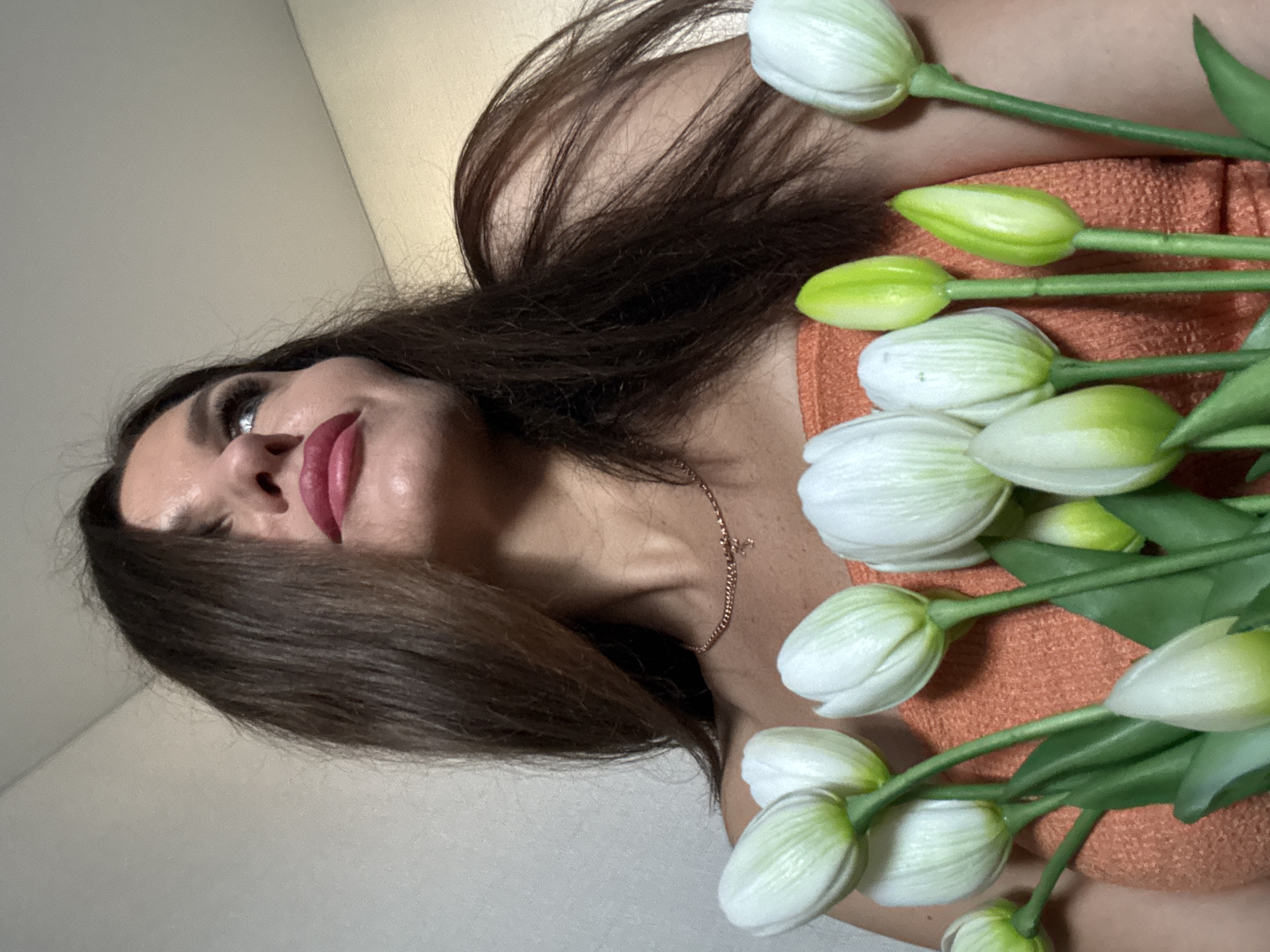 A woman with long, brown hair is lying down and holding a bunch of white tulips. She is wearing an orange top and a gold necklace. The background is a plain, light-colored wall. The image was taken in Chaikovsky, Russia.