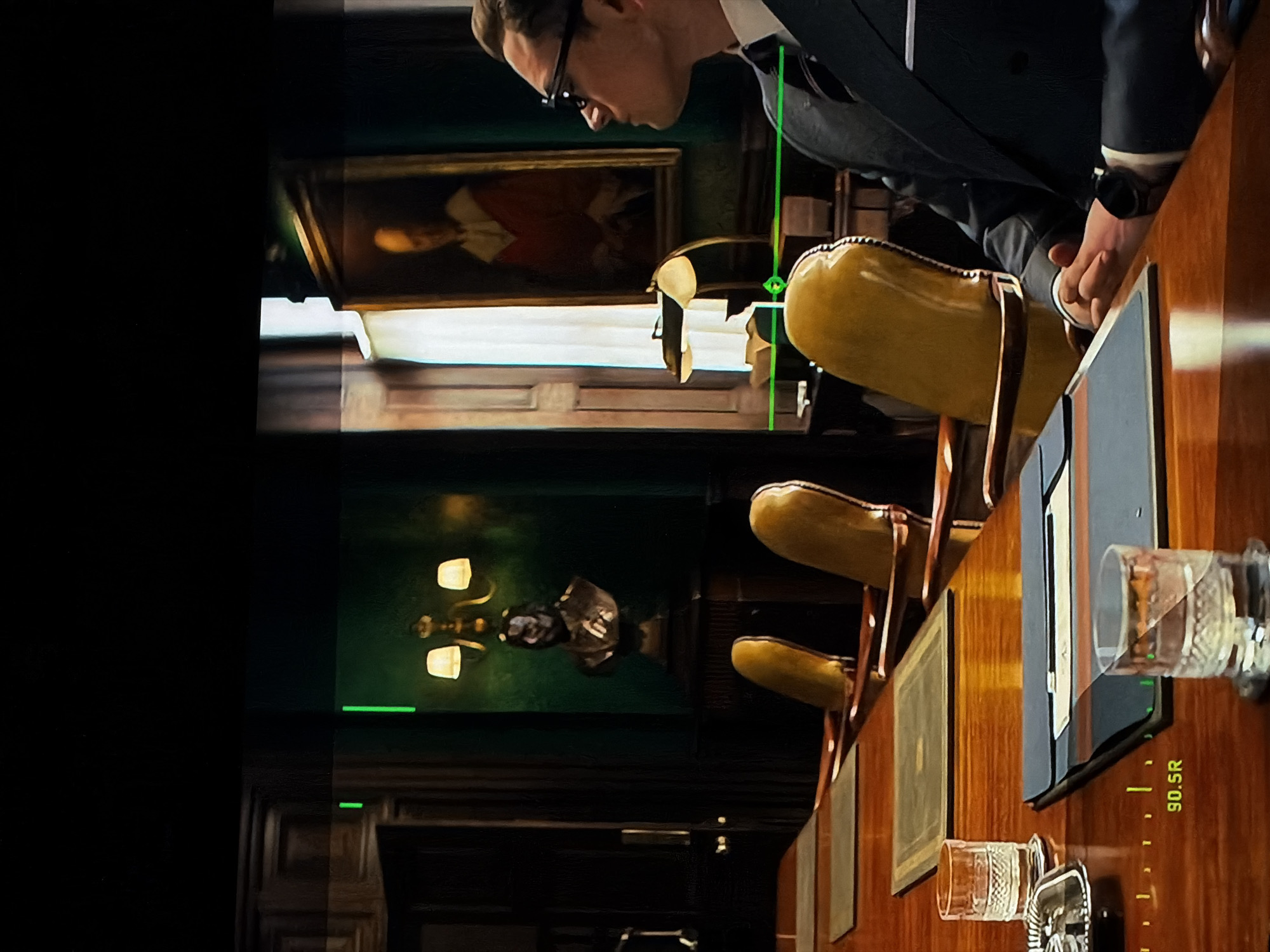 The image depicts an indoor scene, likely a formal study, office, or meeting room, characterized by rich, dark wood paneling and dark green walls. The setting features traditional decor, including a large, highly polished wooden table and several ornate chairs with bright yellow/gold upholstered backs and dark wood frames.
The primary subject is a man, partially visible, leaning over the wooden table. He wears dark-rimmed glasses, a dark suit jacket, a light-colored shirt, and a tie. His left hand, adorned with a dark wristwatch, rests on the table next to a dark, rectangular object that resembles a monitor or tablet. His gaze is directed downwards, suggesting focus on an item on the table.
On the table, two ornate, possibly cut-glass, drinking glasses are present. One appears empty, while the other might contain liquid or be reflecting light. The polished surface of the table reflects elements of the room, including the chairs and glasses.
The background features a framed painting on the dark green wall, depicting a portrait of a person in historical attire, possibly a red coat. A wall-mounted light fixture with two glowing, bell-shaped lamps provides warm illumination further in the background.
Several bright green lines are overlaid on the image, appearing as digital indicators or an interface element. One prominent vertical green line extends from near the man's chest towards the table, intersecting with a small green circular crosshair. Another shorter horizontal green line is visible on the wall. The text "90.5R" is visible on the wooden table surface, near one of the glasses. The overall atmosphere is formal and somewhat contemplative.