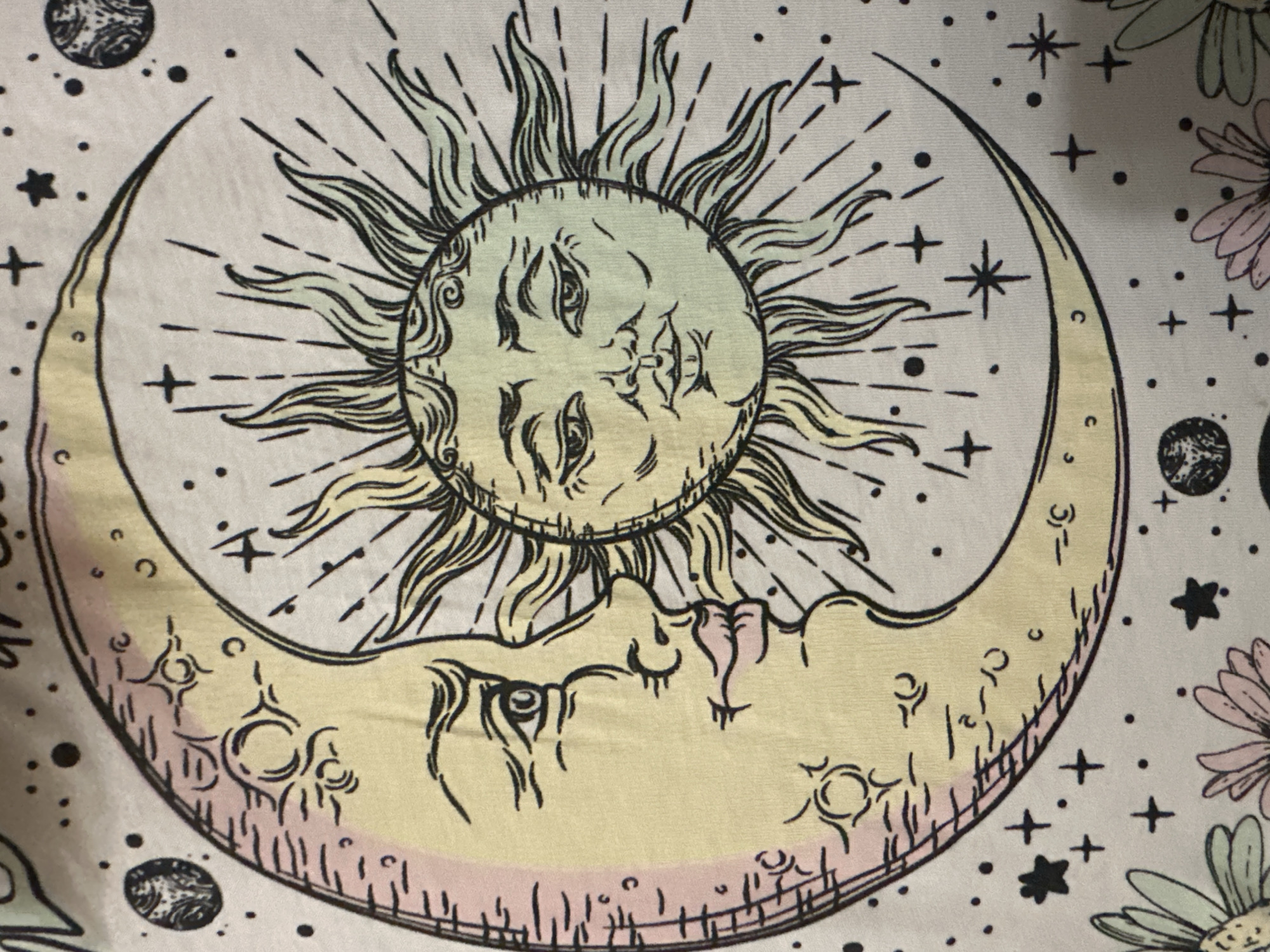 The image displays a close-up of a white fabric with a celestial design. The design features a crescent moon with a human-like face on the left and a sun with a human-like face on the right, partially overlapping and forming a circular composition. The moon is colored in pale yellow and pink, with black outlines and details. The sun is colored in pale yellow and light green, also with black outlines and wavy rays. The background is dotted with small black circles and stars, and some larger, detailed planets or celestial bodies. At the bottom of the image, parts of two daisy-like flowers are visible, with pale green and pink petals. The fabric appears to be slightly creased, suggesting it might be a piece of clothing or a tapestry. The image is a static, two-dimensional representation of this design. The location is unknown.
