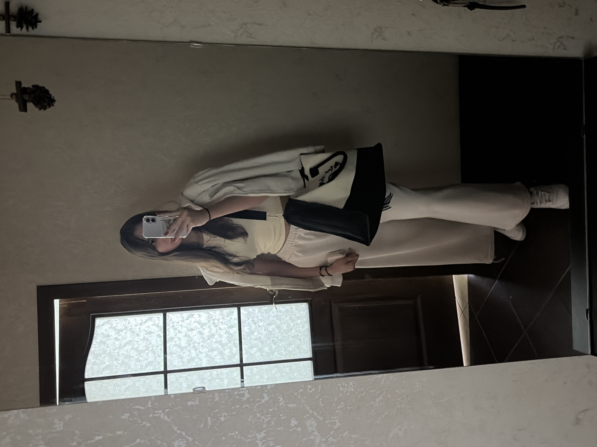 A young woman is taking a mirror selfie in a hallway. She is wearing a cream-colored crop top, matching wide-leg pants, a light beige jacket draped over her shoulders, and white sneakers. She is holding a smartphone in her right hand, and a tote bag with black and white panels is slung over her left shoulder. The hallway features a dark brown door with a frosted glass panel to her left and a large mirror on the right wall. Two dark, decorative objects hang from the wall above and behind her. The flooring appears to be dark tiles. The overall lighting is dim. This scene could be located within a residential building in Tiraspol, Moldova.