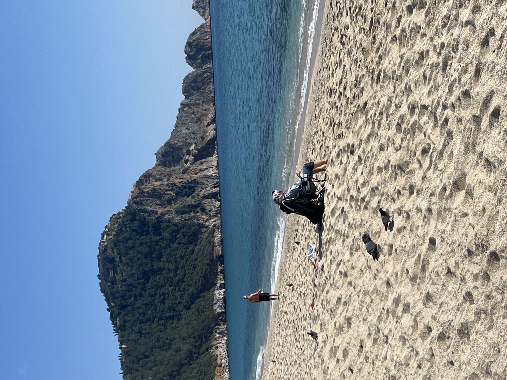 The image displays a daytime scene on a sandy beach in Alanya, Turkey, under a clear blue sky. The prominent background features a large, densely forested hill topped by the fortified walls of Alanya Castle on the left, transitioning into rugged rocky cliffs and smaller islets extending into the blue-green sea on the right.
In the mid-ground, a lone individual sits facing the sea in a dark folding beach chair on the light brown sand. This person is wearing dark long pants and a light-colored head covering, with a dark backpack positioned next to the chair. A red and white striped fabric item is spread on the sand nearby.
To the left of the seated individual, closer to the water's edge, a male figure stands with his back to the viewer, facing the sea. He is shirtless and wears dark shorts, holding what appears to be a fishing rod in his right hand. Gentle waves are visible breaking on the shore. Scattered across the sandy foreground are several pigeons. The sand shows numerous footprints.
