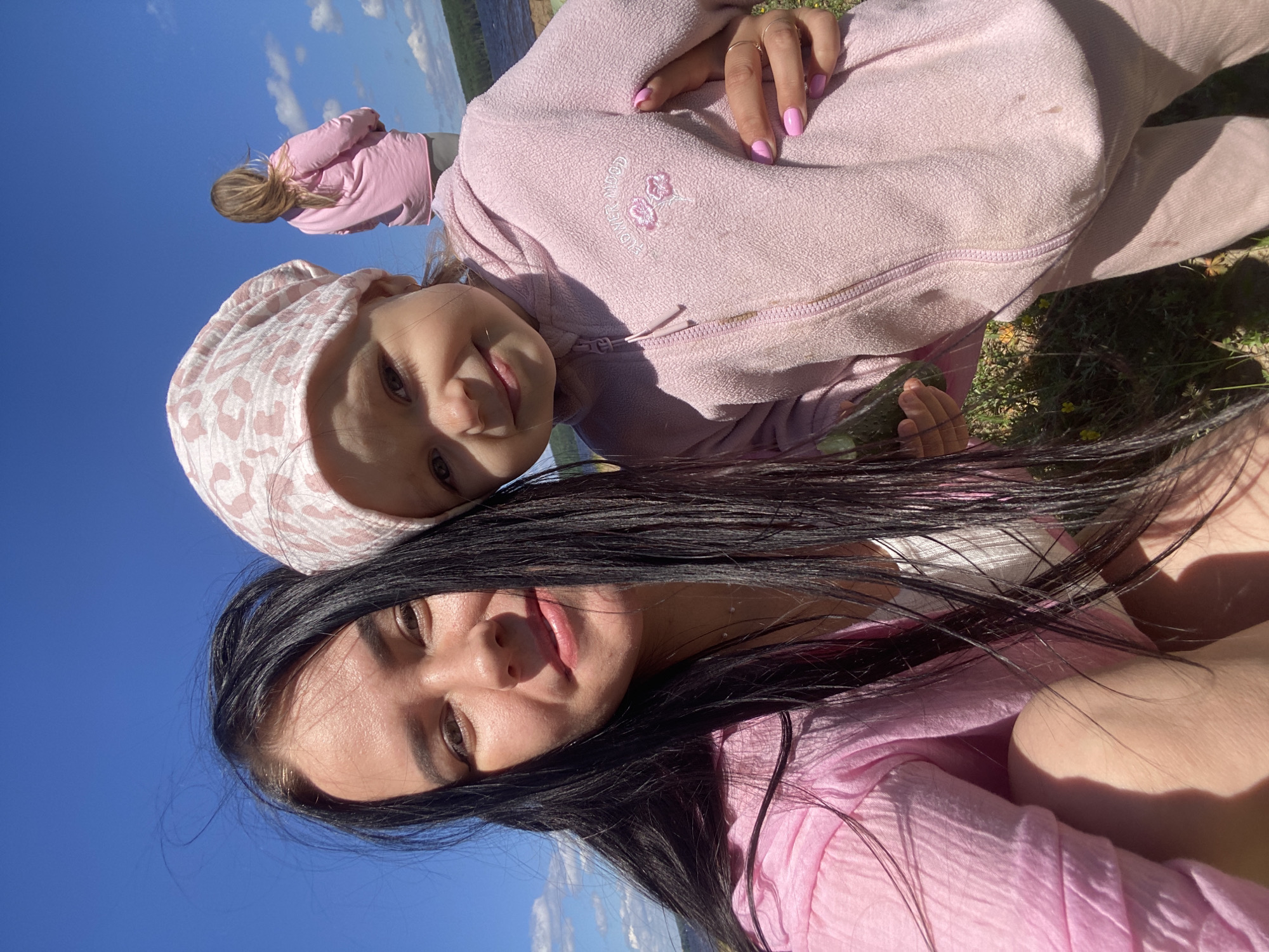 The image shows three individuals outdoors on a sunny day, likely in Chaikovsky, Russia. The primary focus is on a woman in the foreground and a young child positioned above her. Both are wearing pink clothing. The woman has long, dark hair and is looking towards the camera with a relaxed expression. The child, wearing a pink patterned hat, is looking up with a slight smile. Behind them, a third person, also wearing pink, is visible in the distance, with their back to the camera. The background consists of a clear blue sky with scattered clouds and a body of water, suggesting a lakeside or riverside location. The overall mood is joyful and familial, indicative of a casual outdoor outing. The light suggests it is daytime. There is no visible text in the image.