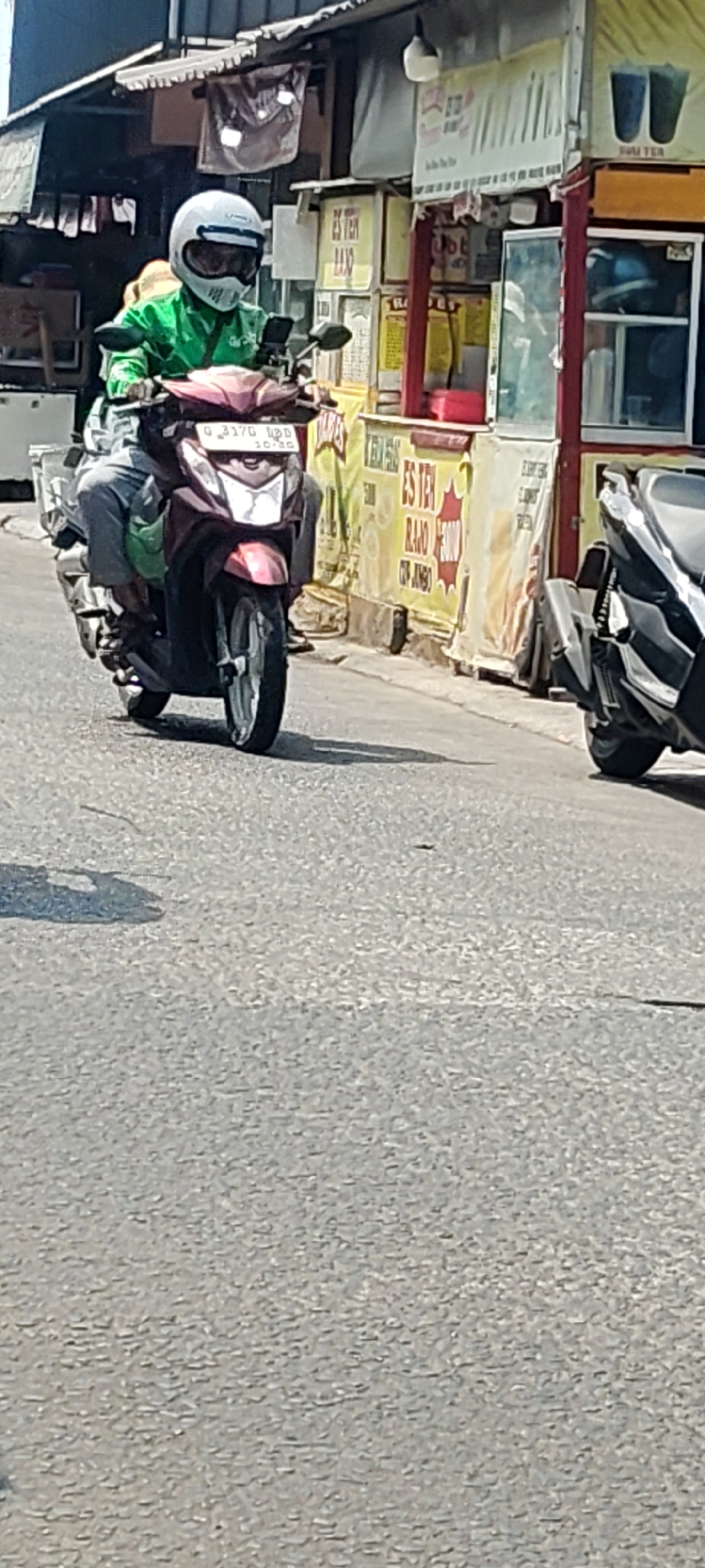 A person wearing a green jacket and a white helmet is riding a maroon motorcycle. The motorcycle has a license plate that reads "G 3170 UDD". In the background, there is a food stall with yellow banners that have Indonesian text on them. The stall appears to be selling drinks, with images of cups and text that says "ES TEH RAJO" and "JUMBO". There are other motorcycles parked in front of the stall. The scene is taking place on a paved street in what appears to be a commercial area. The lighting suggests it is daytime. The overall impression is a busy street scene in a city. The image is grainy and out of focus in some areas. Based on visual cues like the architecture of the stall and the style of the motorcycles, this scene is likely in Indonesia, possibly in Mekarmukti, as indicated by the provided context.