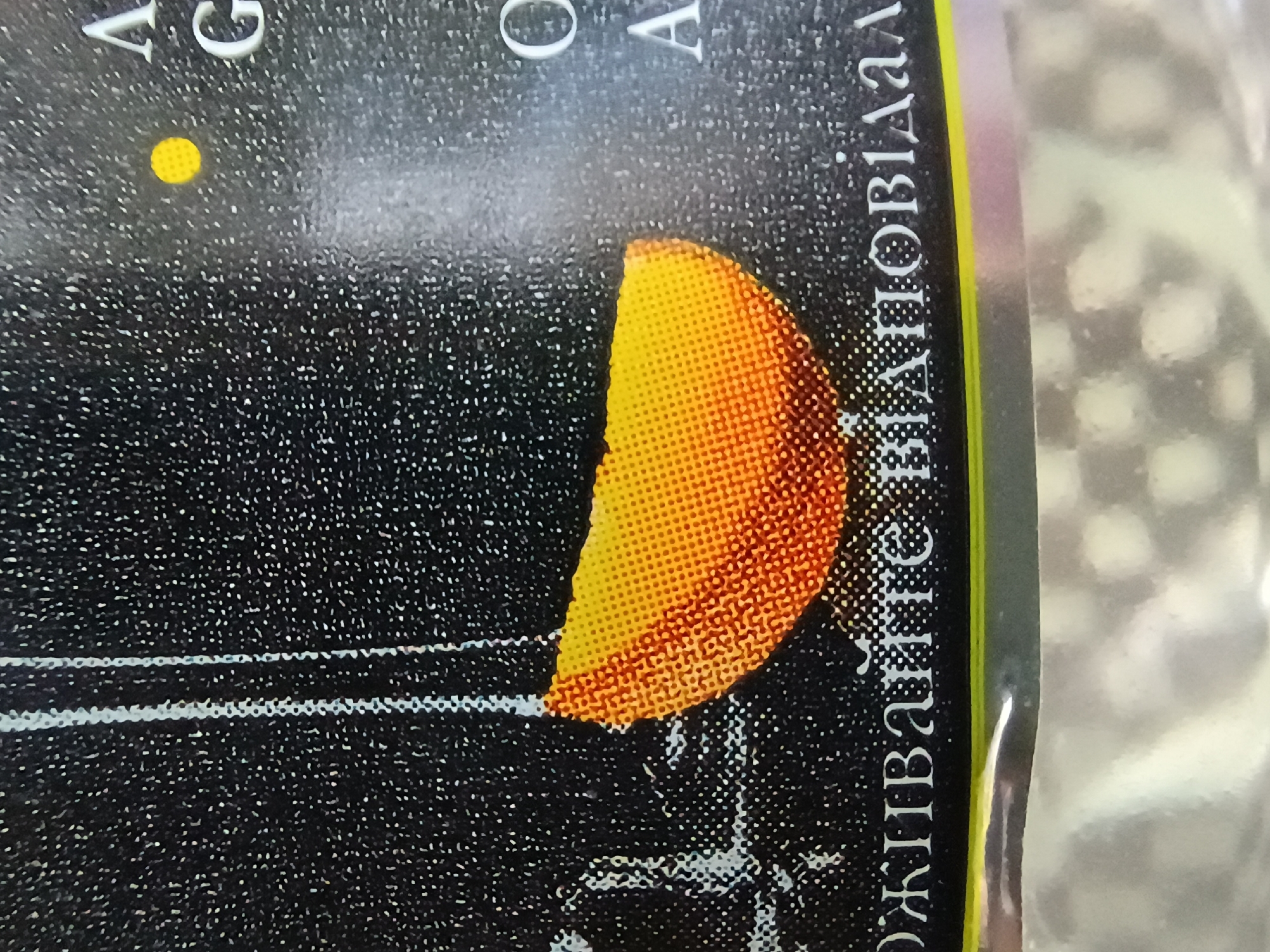 This image shows a close-up of a book cover, likely a paperback given the flexible appearance. The cover has a dark, starry background with a prominent crescent-shaped object that appears to be a celestial body, possibly the moon or a stylized sun, rendered in shades of orange and yellow. The object is detailed with a stippled texture.
There is text visible on the cover, running vertically along the right side of the image. The text is white and appears to be in a Cyrillic alphabet. Some legible characters include "А", "Г", "О", and "Ж". The words seem to be a title or author's name. In the upper left corner, a small yellow circular dot is visible, potentially representing another star or planet. Two thin, horizontal white lines are also present in the lower left, suggesting a graphic design element or part of a larger illustration.
The overall impression is cosmic or astronomical. The lighting is not indicative of a specific time of day, and the setting is abstract, limited to the book cover design. No people are visible. The image is likely a detail from a book being sold or displayed in Zaporizhzhia, Ukraine, as indicated by the provided location context.