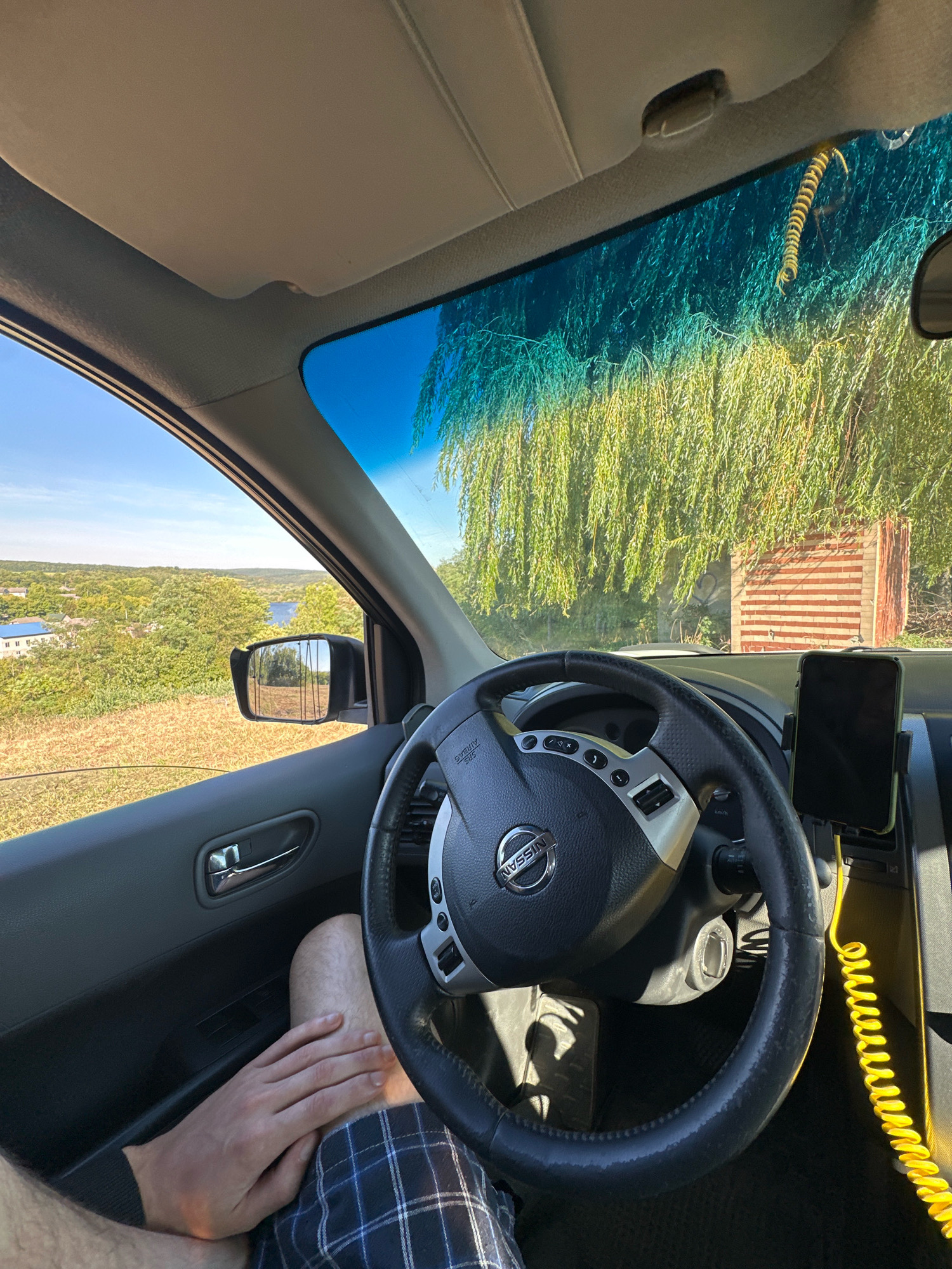 The image provides an interior view from the driver's seat of a vehicle, looking out the windshield and side window. The scene is set outdoors, with a scenic landscape visible through the windows, characterized by rolling hills, scattered houses with blue roofs, dense green trees, and a body of water in the distance. The time of day appears to be sunny, with a clear blue sky.
Dominating the foreground is the steering wheel of a Nissan vehicle, with various controls on its spokes. A smartphone is mounted on a holder to the right of the steering wheel. A coiled yellow cord is visible, likely a charger or cable, extending from the phone mount downwards. The driver's legs and left hand are visible in the lower portion of the frame; the hand rests on the driver's thigh, which is covered by plaid shorts. The interior of the car is primarily dark gray and black, with a beige headliner and sun visor.
Outside the driver's side window, a side-view mirror reflects a rural landscape. The windshield reveals a large tree with long, drooping branches, appearing to be a willow, and behind it, a rustic structure made of red and white striped bricks.
The location is specified as Makiv, Ukraine. The image captures a moment of pause or journey, with the driver at the helm, looking out at the picturesque Ukrainian countryside.