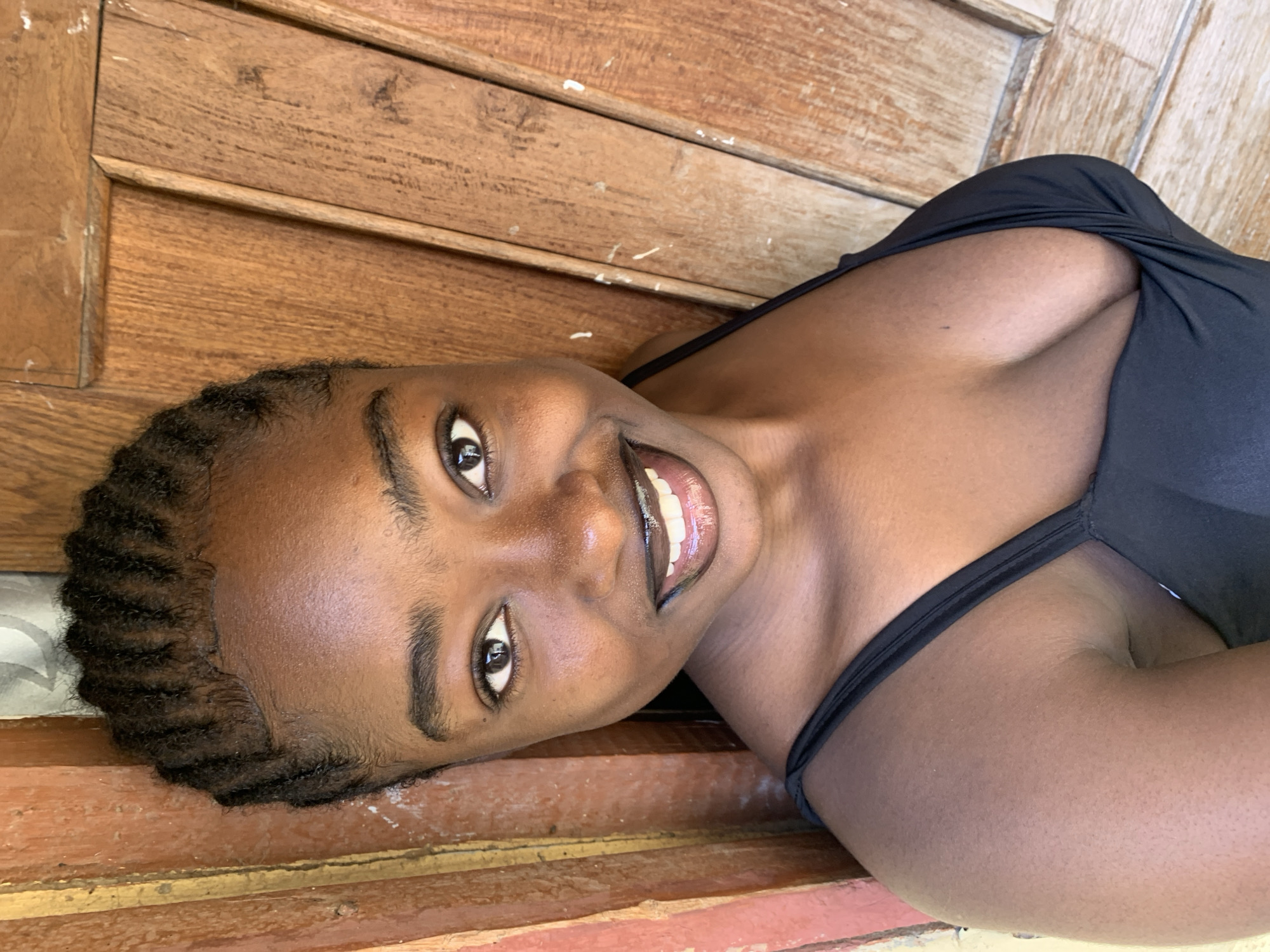 This media file is a portrait of a young Black woman lying down on what appears to be wooden planks. She is wearing a black, low-cut top. Her head is tilted, and she is looking directly at the camera with a bright smile, revealing white teeth and shiny lips. Her dark hair is styled in braids.
The background consists of textured, weathered wooden planks, suggesting an outdoor setting or a rustic interior. The lighting indicates it is daytime, with natural light illuminating her face and accentuating the shine on her skin. The overall mood is relaxed and joyful, conveyed through her expression and pose. The location is identified as Rafin Par, Nigeria. There is no visible text in the image.
