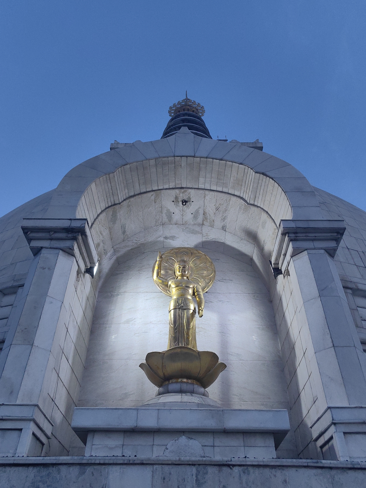The image shows a low-angle, outdoor, daytime photograph of the Vishva Shanti Stupa (World Peace Pagoda) located in Delhi, India. The stupa's white marble dome dominates the upper portion of the frame, with a dark blue sky visible above. A golden statue of Buddha, illuminated by artificial lighting, stands prominently in an arched alcove on the front of the stupa. The Buddha statue is depicted standing on a lotus flower, with one hand raised. The structure features multiple levels of white marble construction, with geometric patterns and architectural details. The lighting suggests it is either early morning or late evening.