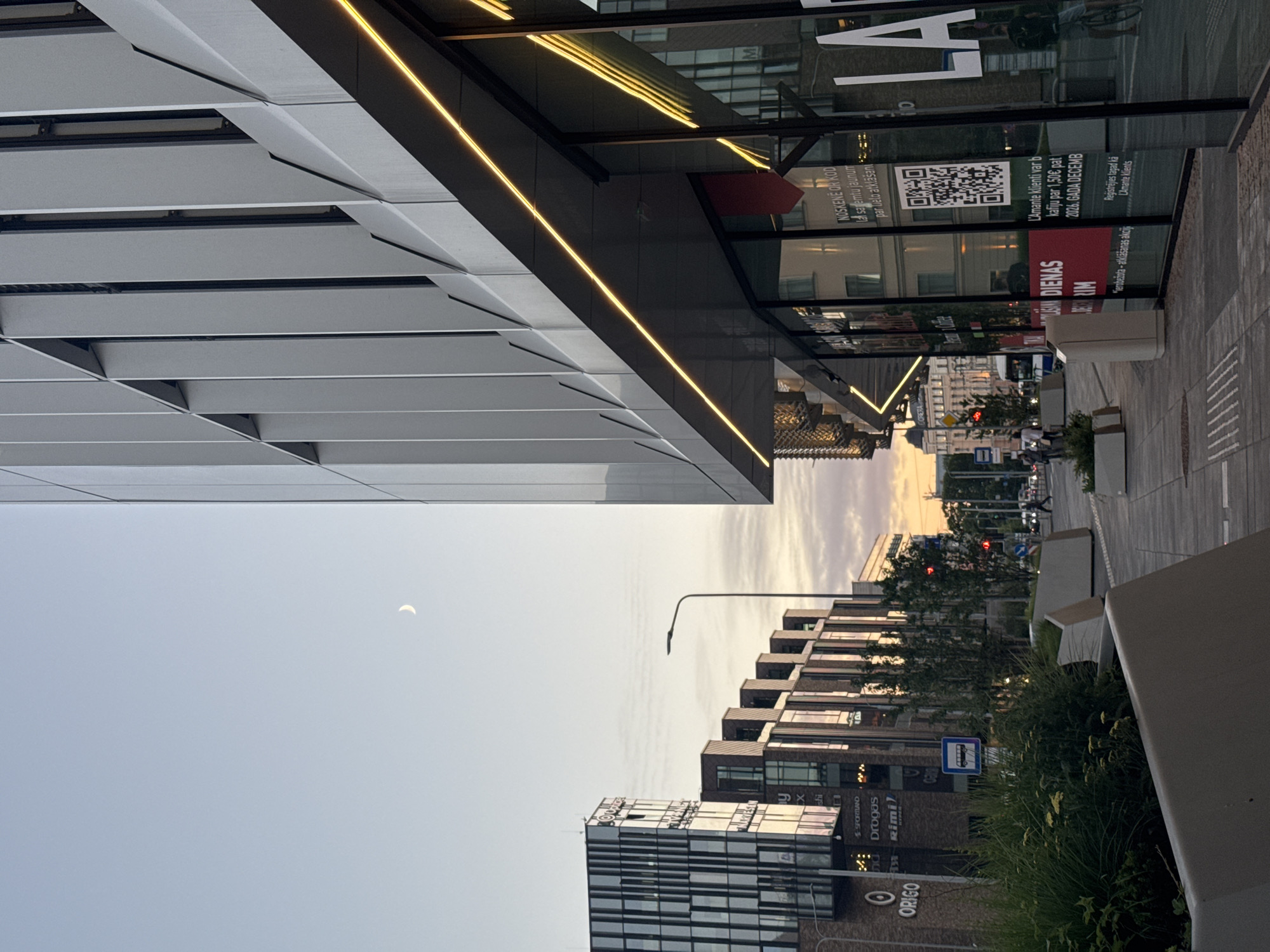 The image depicts a street scene in Riga, Latvia, as twilight descends. A modern, multi-story building with a sleek, metallic facade dominates the right side of the frame, featuring a prominent yellow LED light strip running along its overhang. To the left, a row of newer buildings with glass and brick exteriors are visible, including a building with the "ORIGO" logo. In the sky, a sliver of the crescent moon is present. The sidewalk is paved with light-colored tiles and bordered by planters containing lush green vegetation. A bus stop sign is visible near the center of the image. Further down the street, traffic lights and vehicles can be seen. In the foreground, a person is riding a bicycle past the building. The overall atmosphere is urban and modern, with a touch of natural elements.