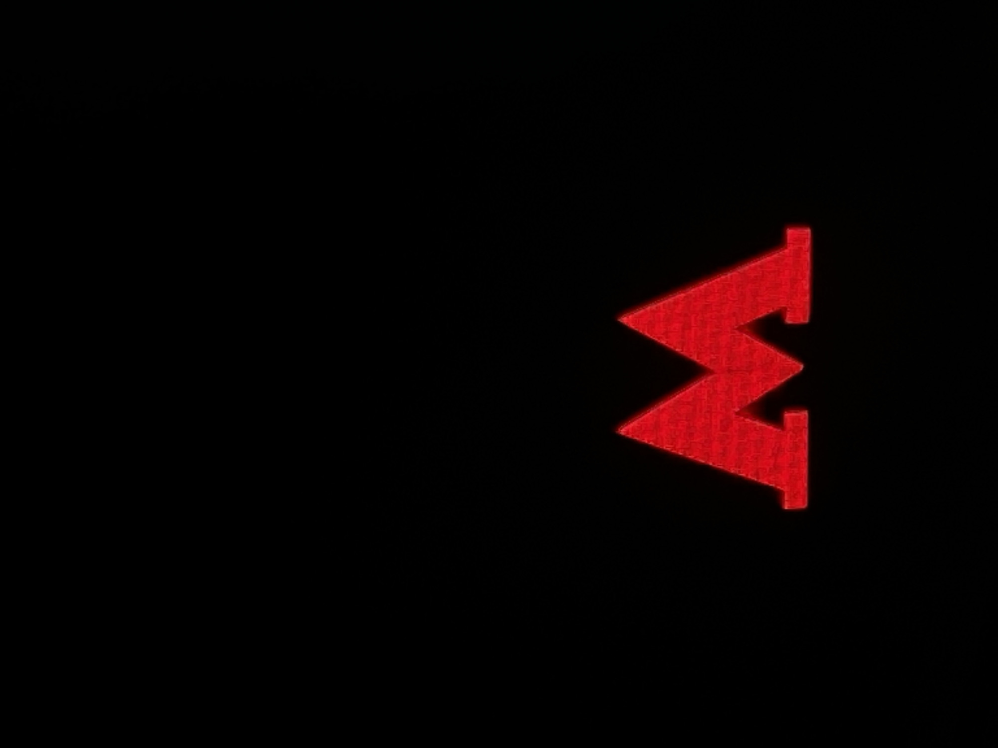 The image displays a prominent red, angular symbol set against a completely black background that occupies most of the frame. The symbol is located in the right half of the image and features a distinct zigzag pattern oriented vertically. It presents two peaks pointing towards the right side of the frame and one trough pointing towards the left. This stylized shape could be interpreted as a letter 'M' rotated 90 degrees clockwise or a 'W' rotated 90 degrees counter-clockwise. The symbol has a textured surface, resembling a rough fabric, pixelation, or embroidery, and subtle shading gives it a three-dimensional effect.
There are no people or other objects visible in the image. No specific activity or event is taking place; the image is a static representation of the symbol. Due to the solid black background, there are no notable details regarding the time of day, weather, or specific location cues within Moscow, Russia. No emotions are conveyed. The only visible text is the described red, stylized letter or zigzag symbol. The scene is minimalist, highlighting the stark contrast between the bright red symbol and the dark backdrop.