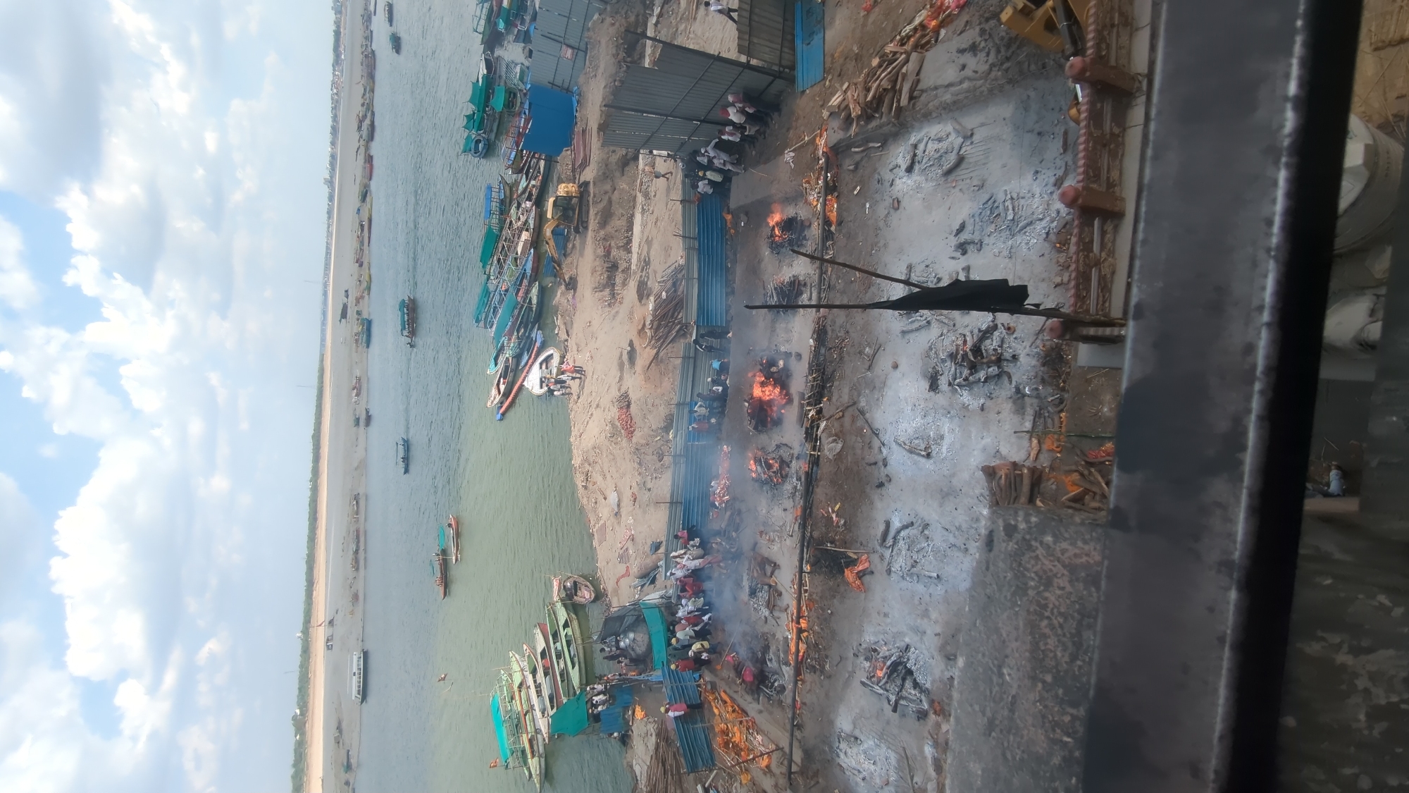 This image captures a view of a cremation ground on the banks of the Ganges River in Varanasi, India. In the foreground, several bodies are being cremated, with visible fires and smoke rising from the pyres. People are gathered around, some tending to the fires, others observing. In the mid-ground, there are piles of firewood and debris. Adjacent to the cremation ground, a fleet of boats is docked along the riverbank, with people visible on some of them. The river itself is wide, with a sandy bank on the opposite side and a distant view of a bridge. The sky above is partly cloudy. The scene depicts a traditional Hindu cremation ritual in progress at a significant spiritual site.