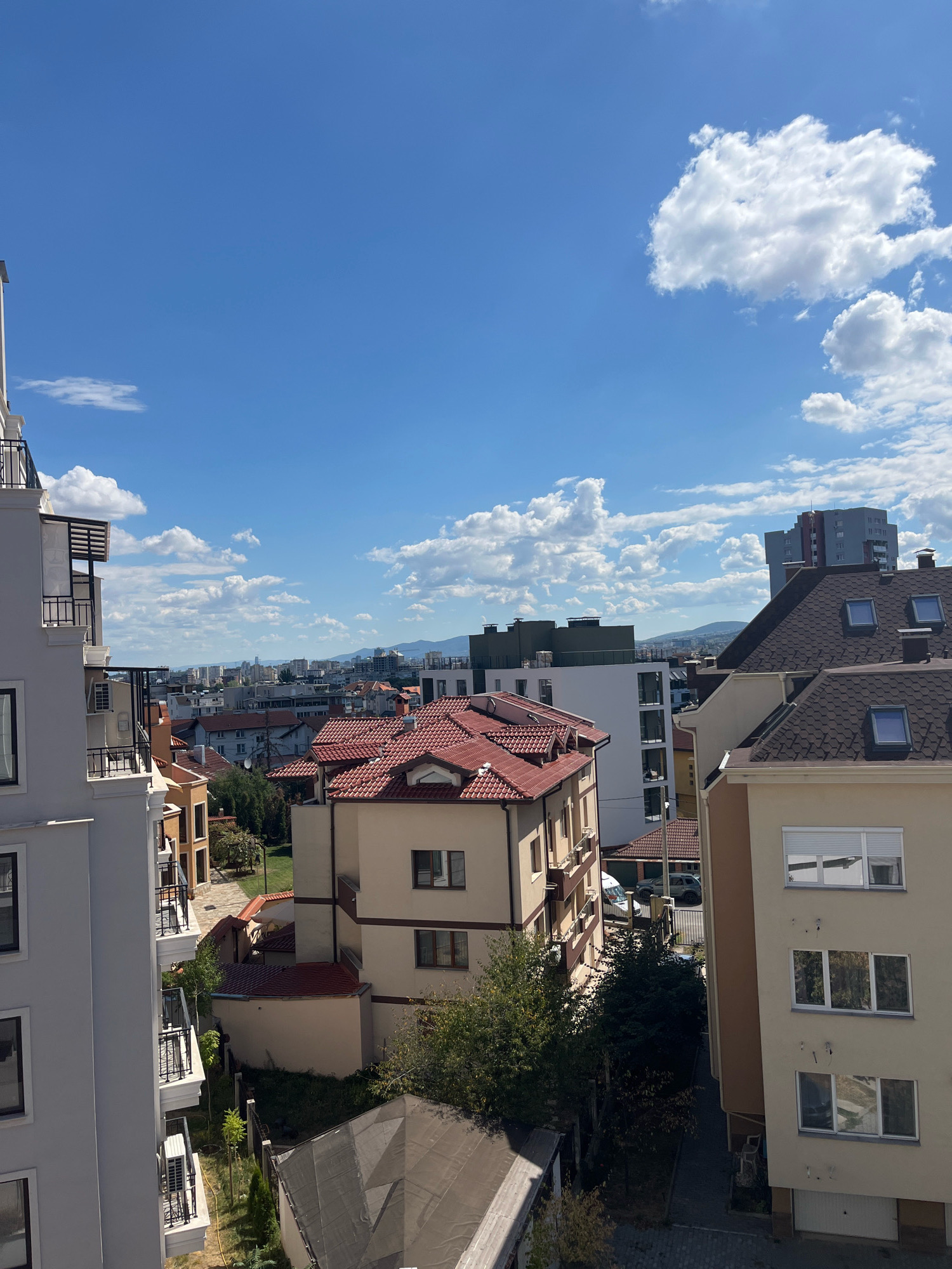 This image depicts a daytime, high-angle view of a residential area in Sofia, Bulgaria. The scene is dominated by various buildings with different architectural styles and roof types, including several with red-tiled roofs and others with darker, slate-like roofing. A prominent modern apartment building with a white facade and balconies is visible on the left. The sky is blue with scattered white clouds, suggesting fair weather. In the background, a cityscape with taller buildings and distant mountains can be seen. The environment appears to be a developed urban area with a mix of residential housing and some commercial structures. There are no people visible in the image. The perspective suggests the photo was taken from a higher vantage point, possibly another building or a balcony.