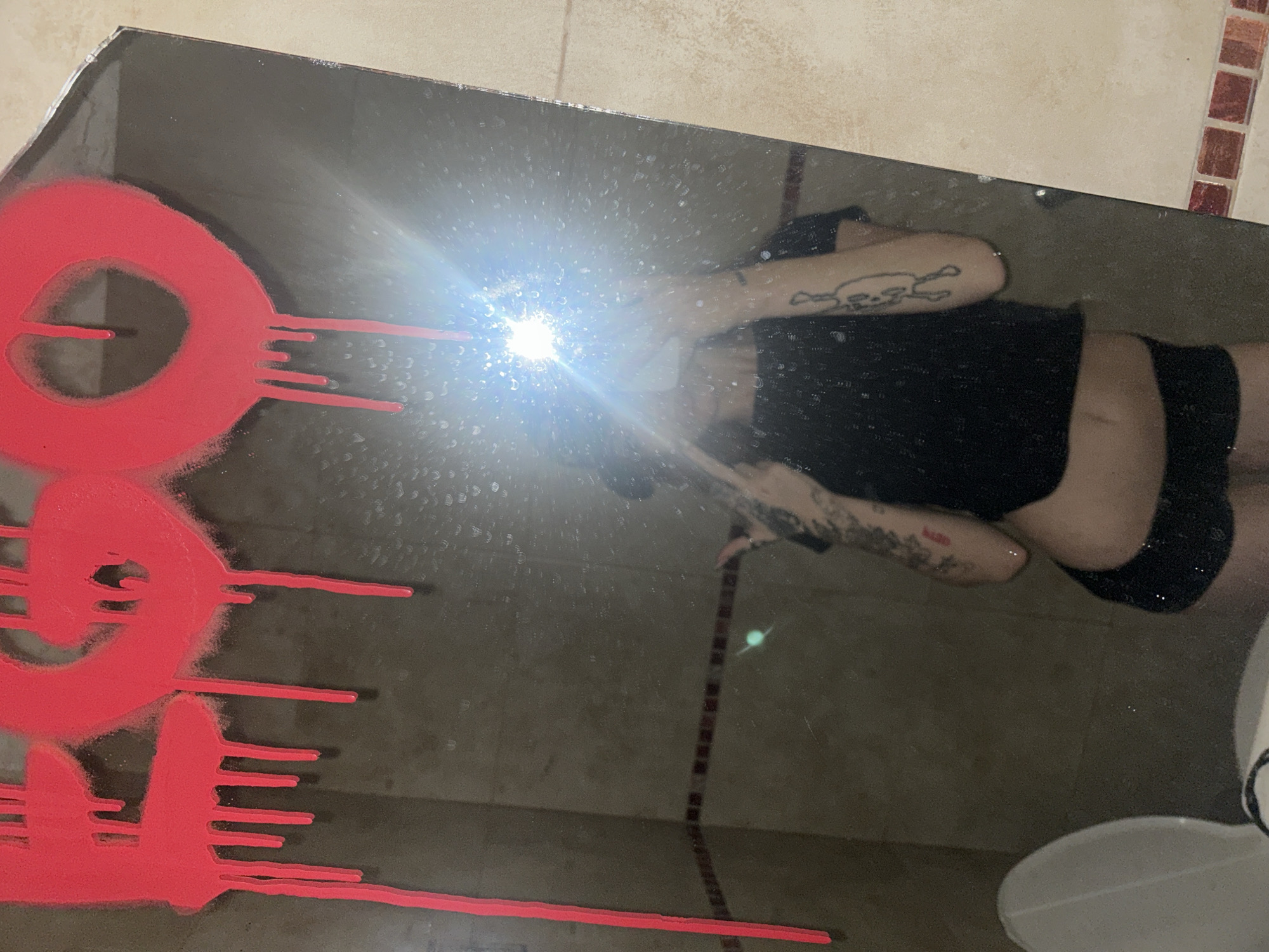 A mirrored surface reflects a person in black attire with tattoos on their arms, holding their hands up to shield their face from the light emanating from the camera flash. Red, graffiti-style markings are visible on the mirror. The reflection also reveals a portion of a bathroom setting, including a toilet and wall tiles. The location is suggested to be Buenos Aires, Argentina, based on the location context of the prompt, although there is no visual confirmation from the image alone.