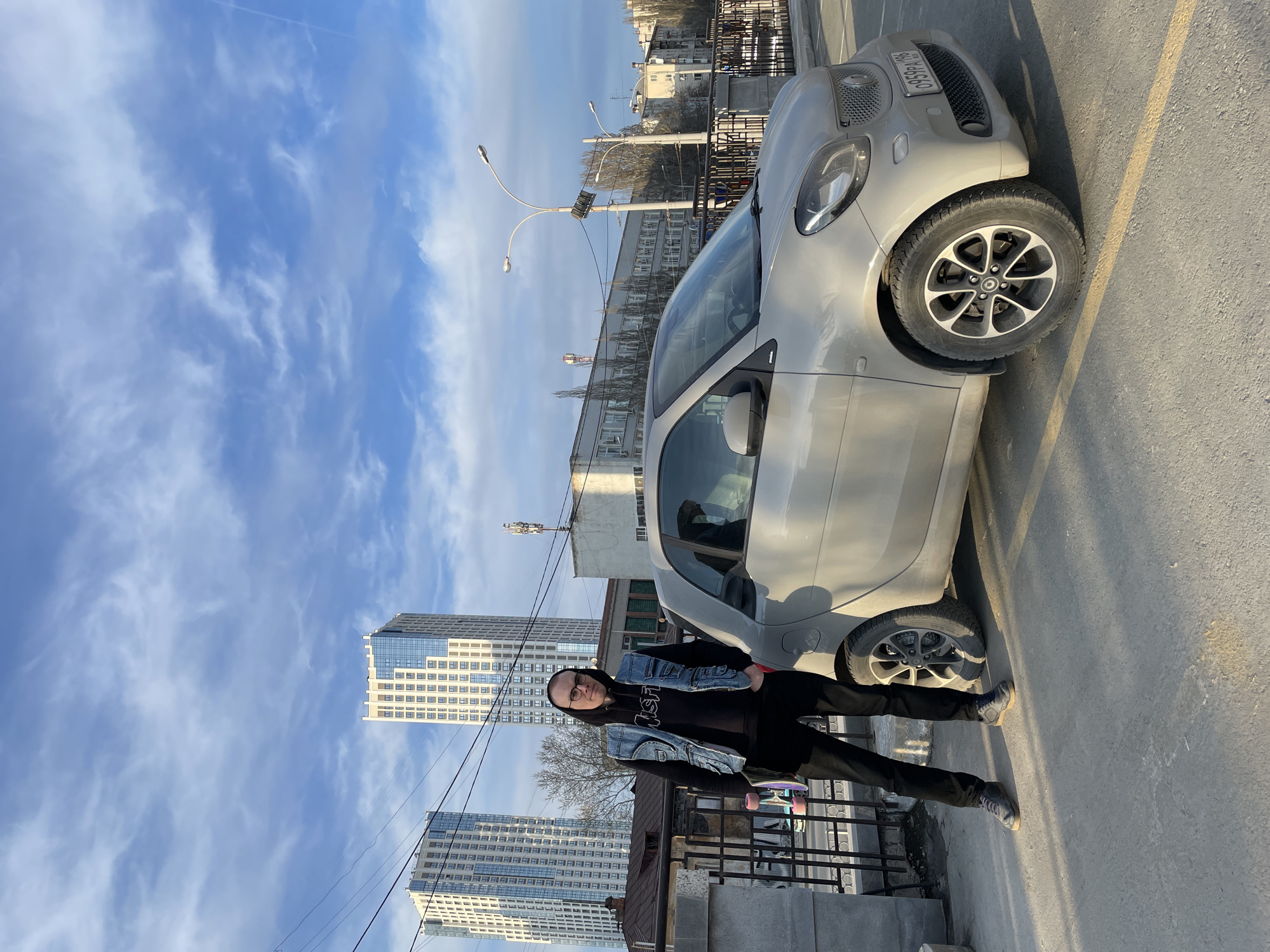 A medium shot shows a man standing next to a silver Smart car. The man is wearing a black hooded sweatshirt, a blue denim vest, black pants, and grey shoes. He holds a skateboard. His glasses are visible. The car is parked diagonally on a grey asphalt surface with yellow lane markings. In the background are two tall buildings, light poles, bare trees, and other structures. The sky is blue with streaks of clouds. The license plate on the car reads "A 684 AB 96". The scene appears to be in an urban area of Yekaterinburg, Russia, as suggested by the architectural style and the license plate format.