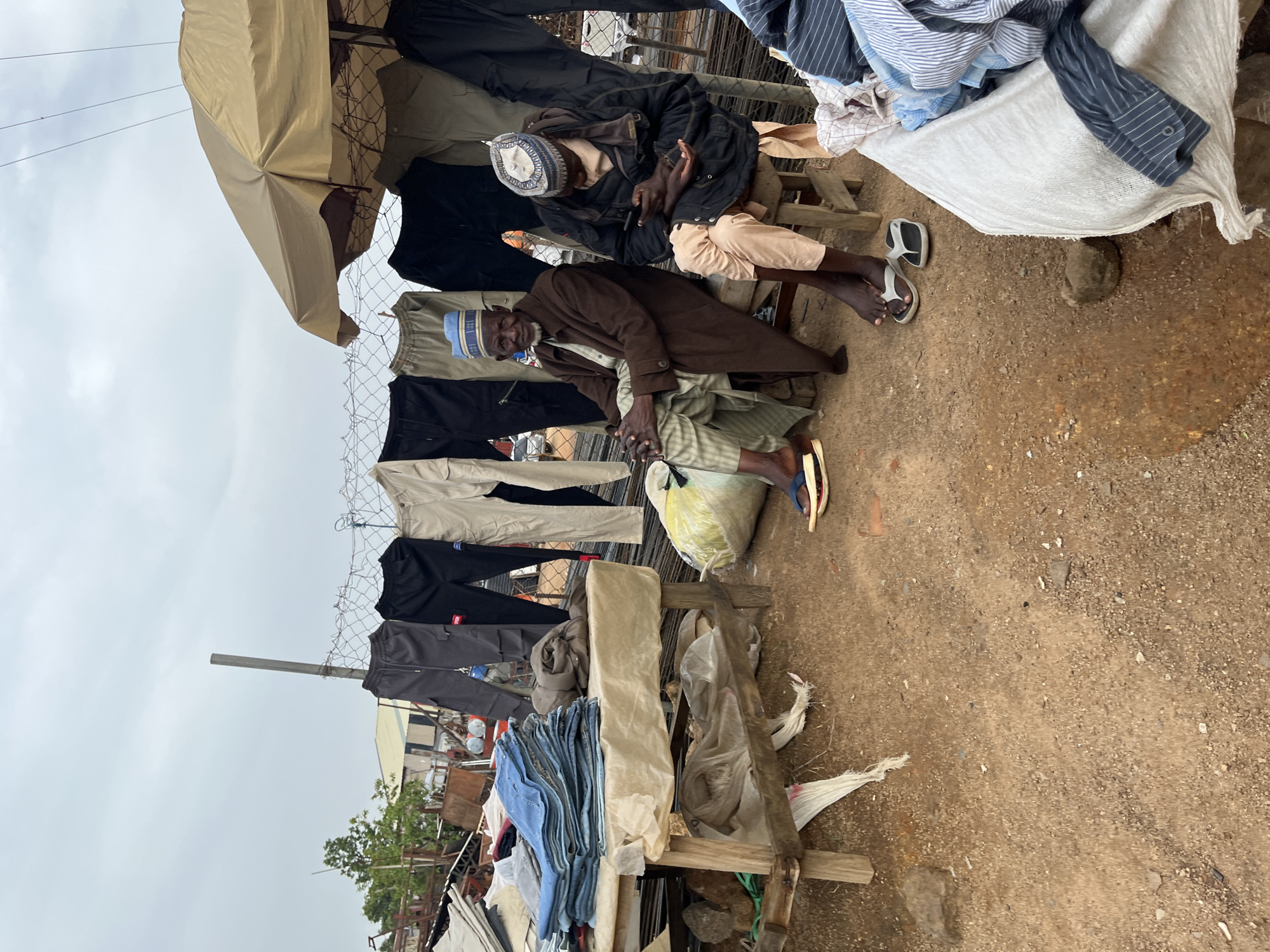 Two elderly men are seated at an outdoor market stall in Gura Top, Nigeria, displaying clothing for sale. The man on the left, wearing a brown coat and a blue and white cap, is looking forward. The man on the right, dressed in a black jacket and a patterned cap, is focused on his mobile phone. Behind them, various trousers and pants are hanging on a wire fence. A table to the left is stacked with folded blue jeans and other garments. A large, yellow plastic bag is on the ground to the right of the seated men, and a pile of striped shirts is visible in the foreground on the right. The ground is sandy and uneven, typical of an outdoor market. An umbrella shades part of the stall. The sky is overcast.